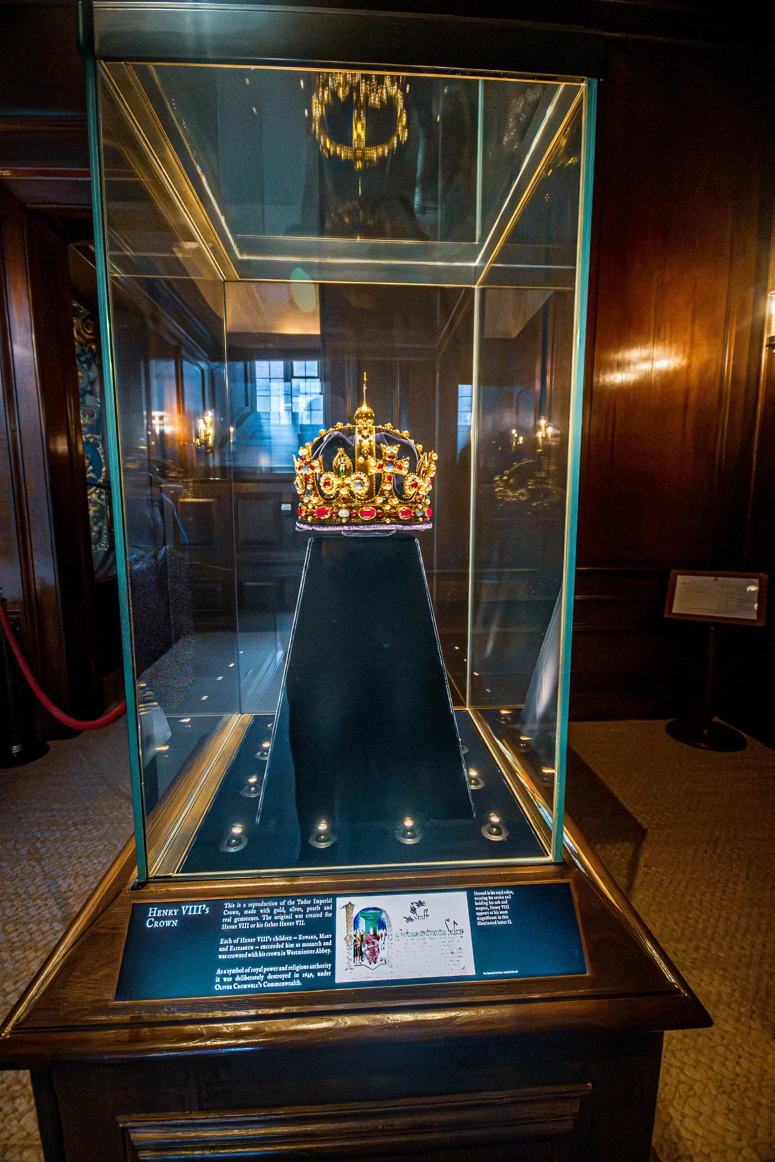 Jeweled crown in a glass museum case on a black pedestal with wooden base and information plaques.