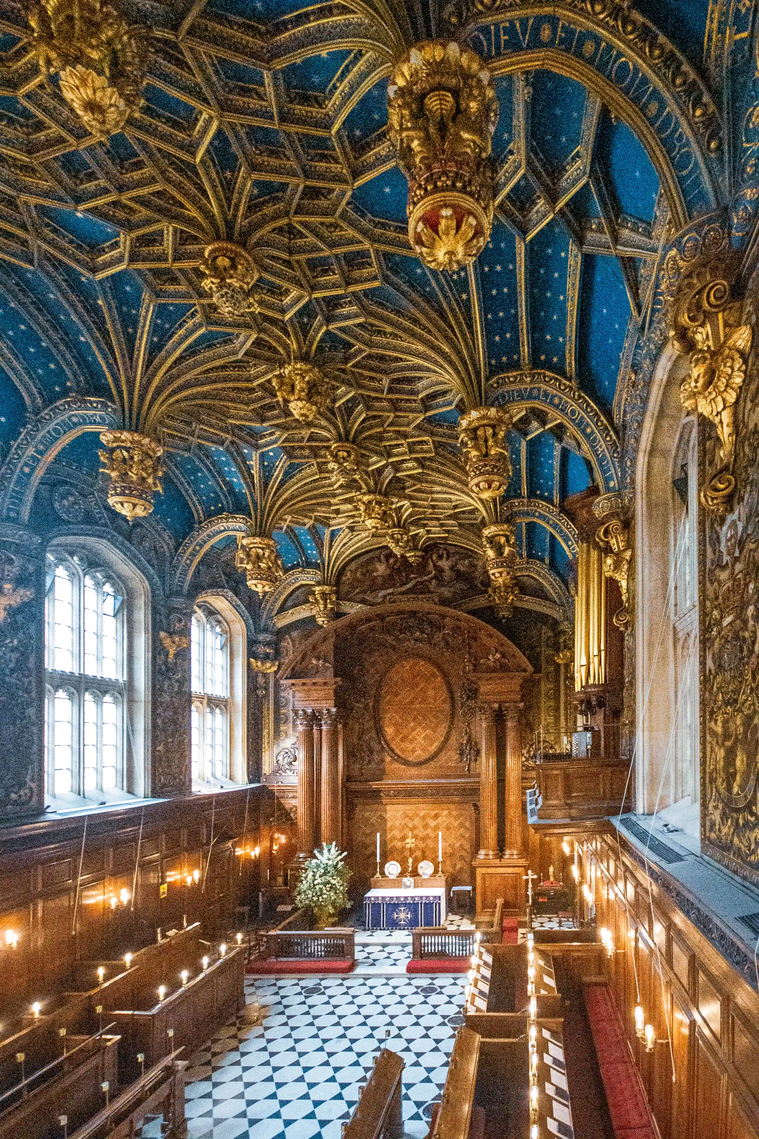Ornate chapel with blue-and-gold fan vaulting, gilded bosses, arched windows, wooden panelling, candle sconces and blue-draped altar.