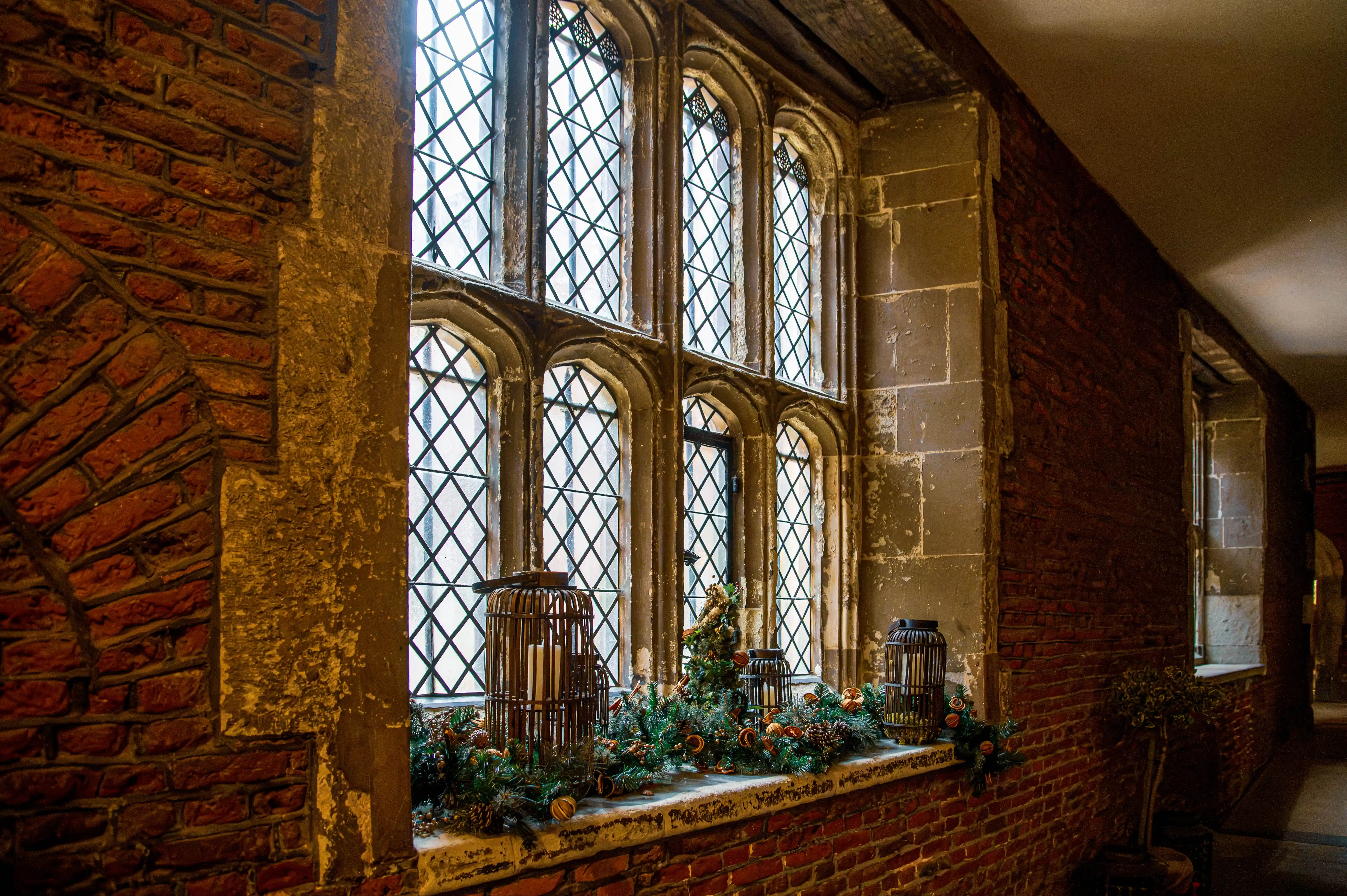 Historic leaded-glass window with festive greenery, pinecones, dried oranges and candle lanterns on a stone sill.