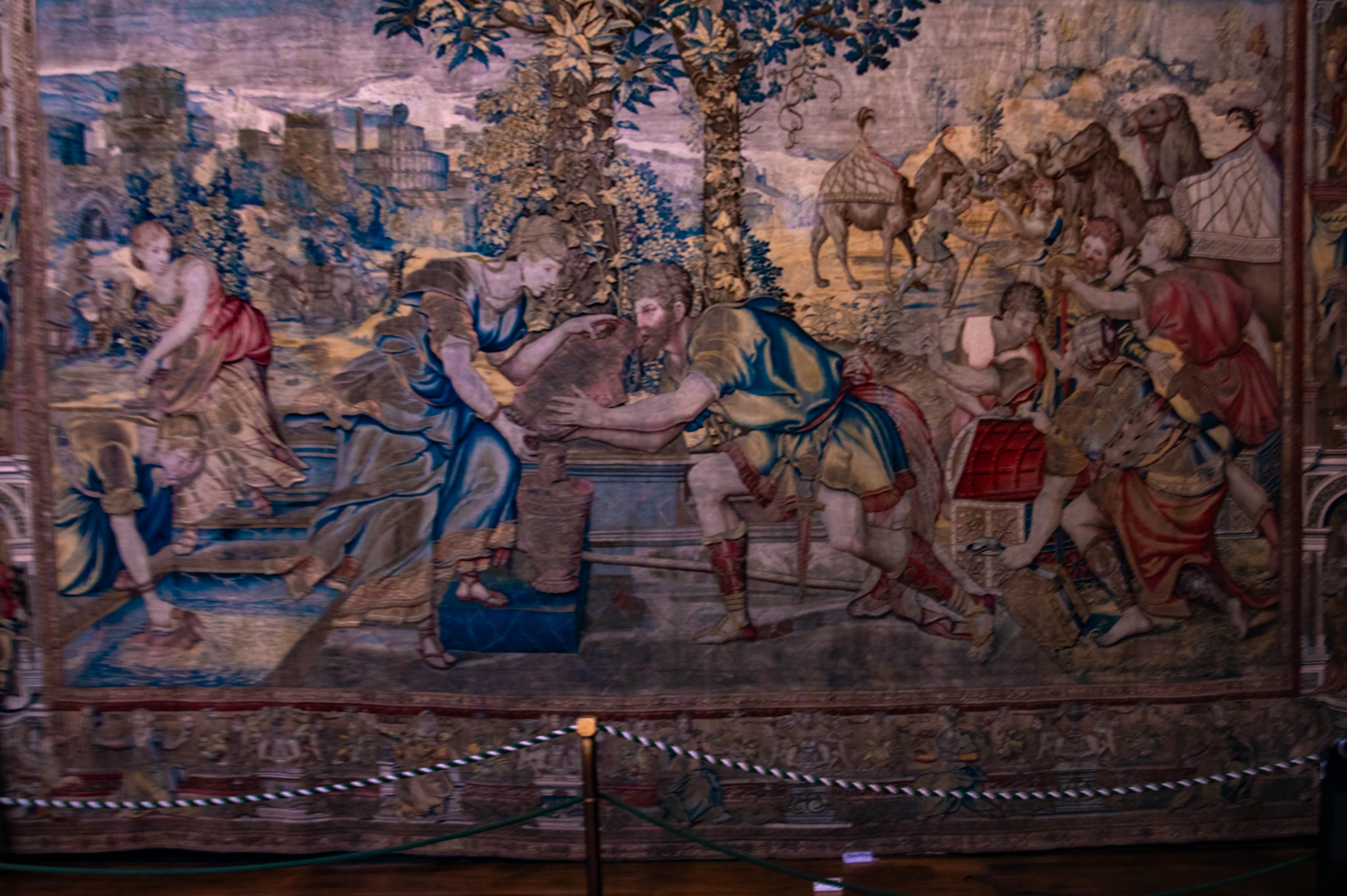 Large historic tapestry showing classical figures, vessels, animals and ancient architectural background, displayed behind a rope barrier.