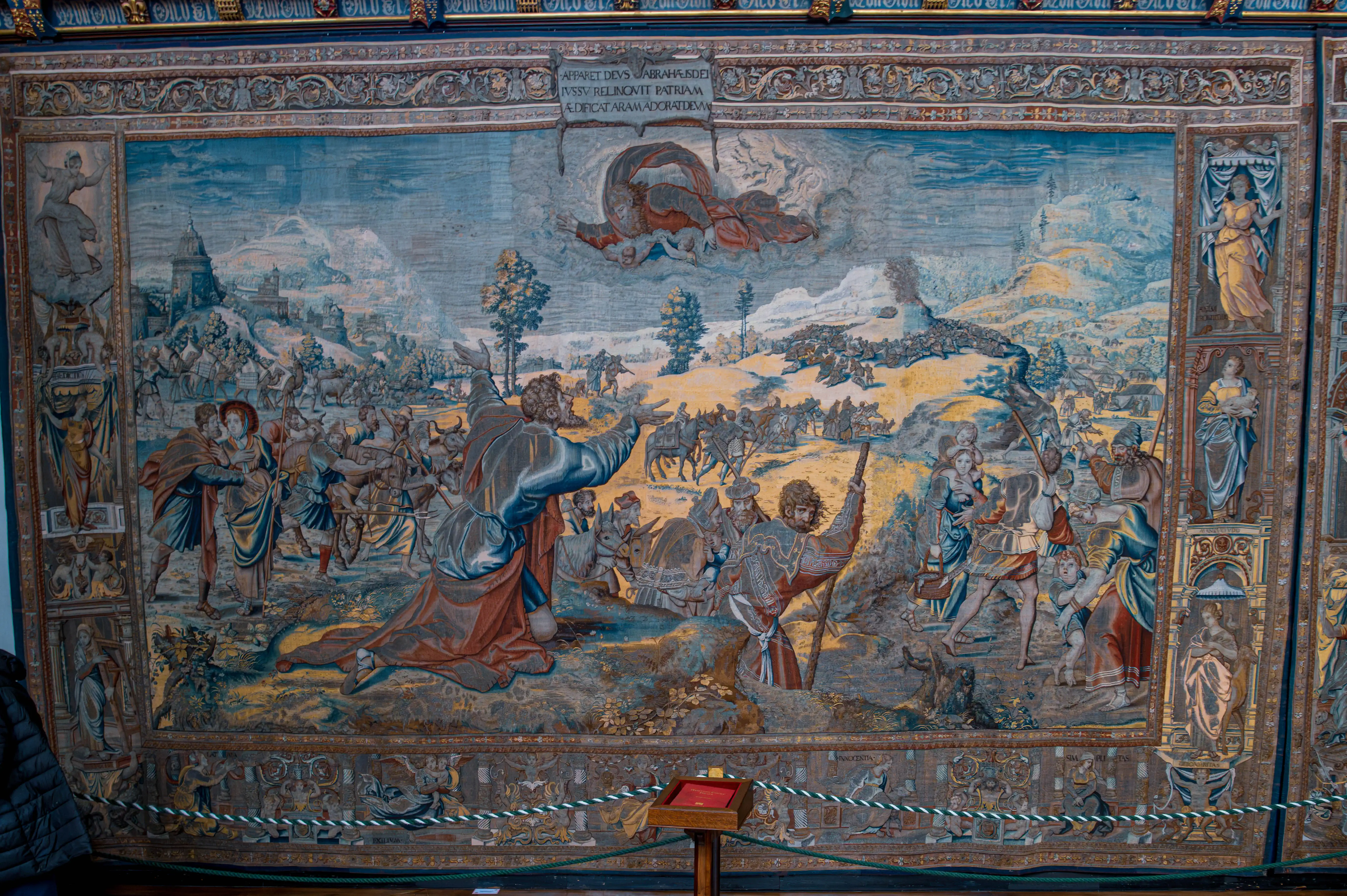 Large historic tapestry showing Abraham kneeling before a divine figure, with surrounding people, animals and ancient landscape, displayed behind a rope barrier.