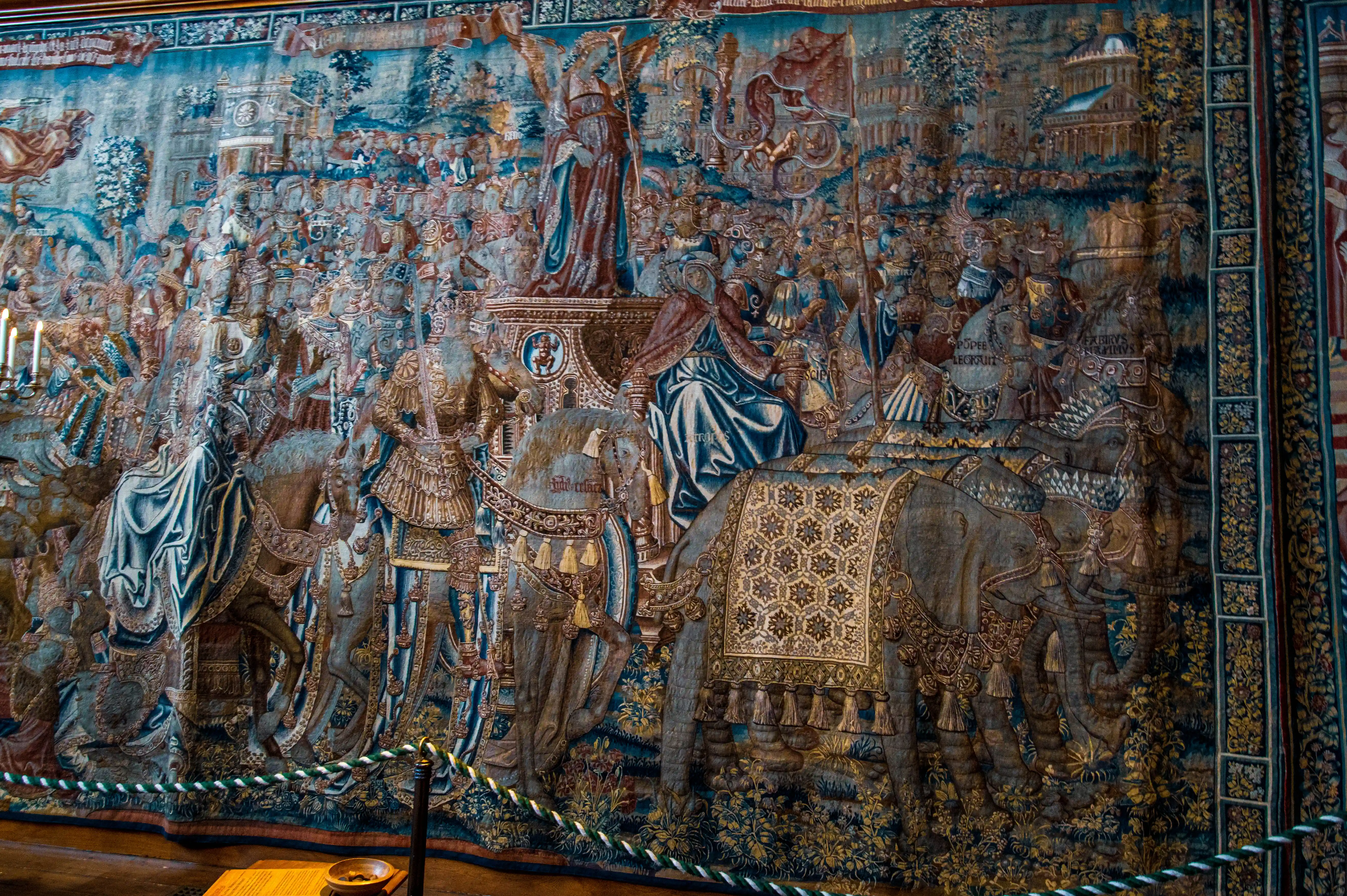 Large historic tapestry showing ceremonial procession with decorated elephants, richly dressed figures and architectural background, displayed behind a rope barrier.