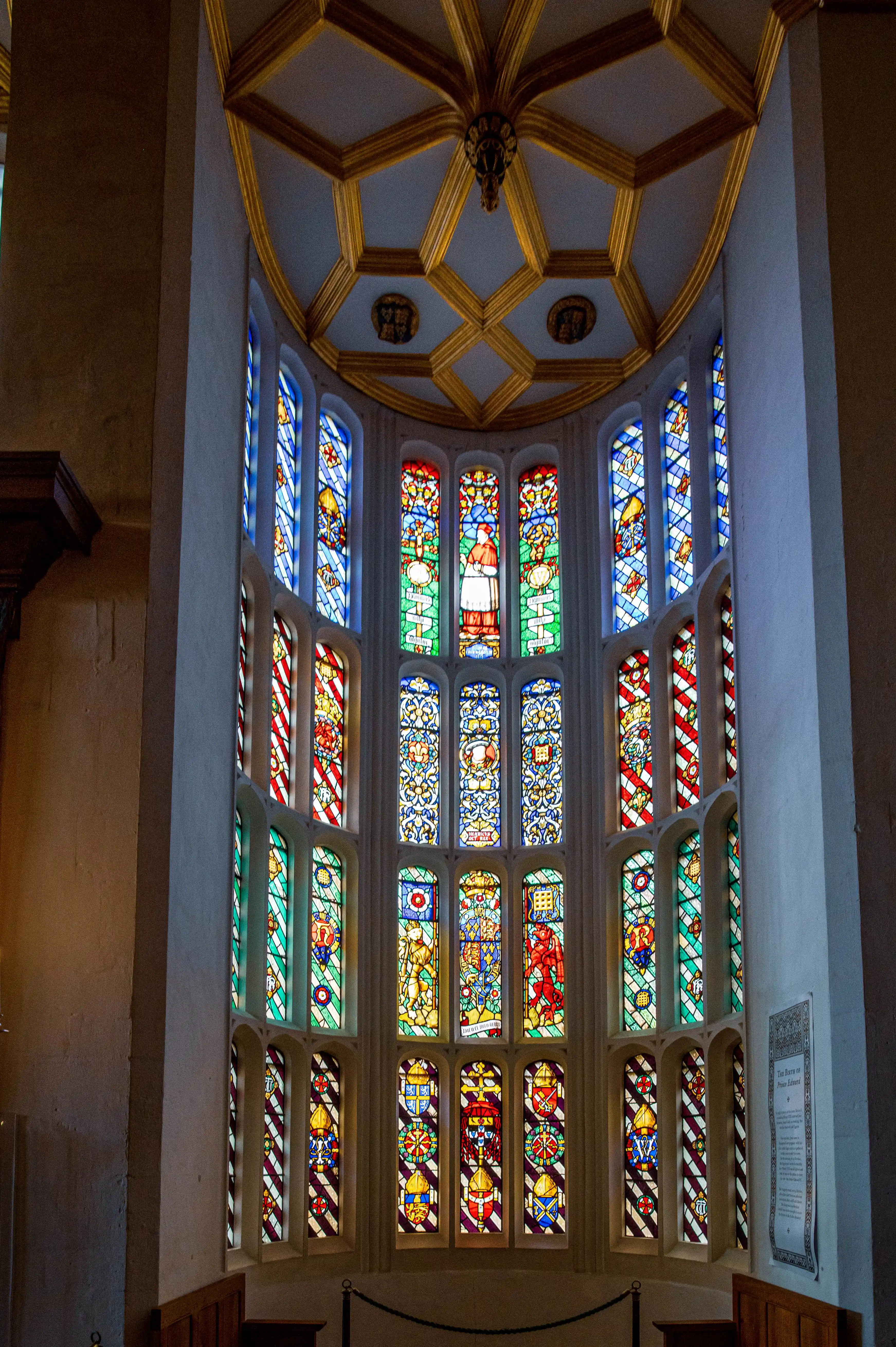 Tall stained-glass window with vivid colours and heraldic motifs beneath a gold-accented geometric ceiling.
