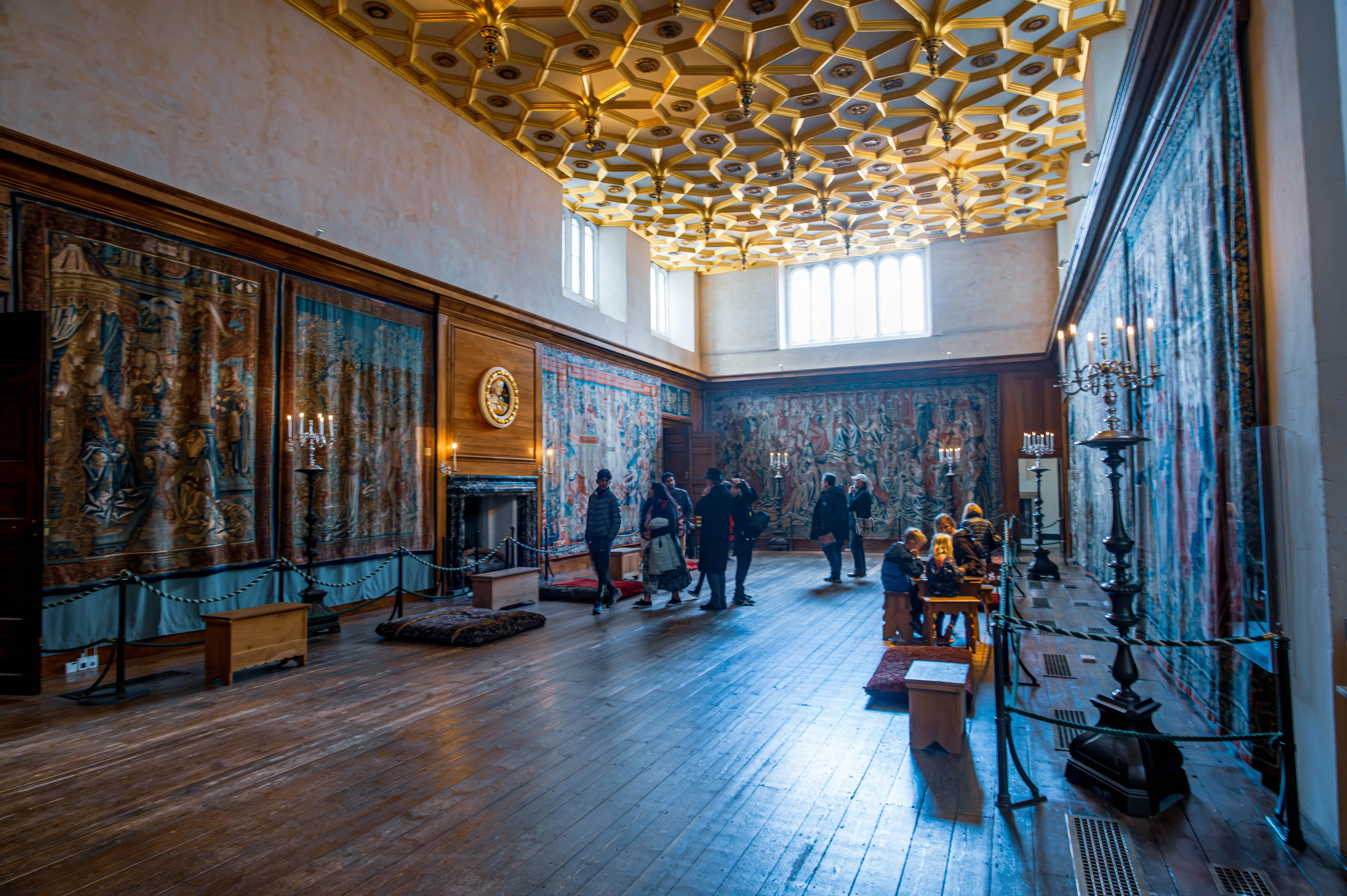 Ornate historic room with large tapestries, gold geometric ceiling, candelabras, fireplace and visitors seated at a wooden table.