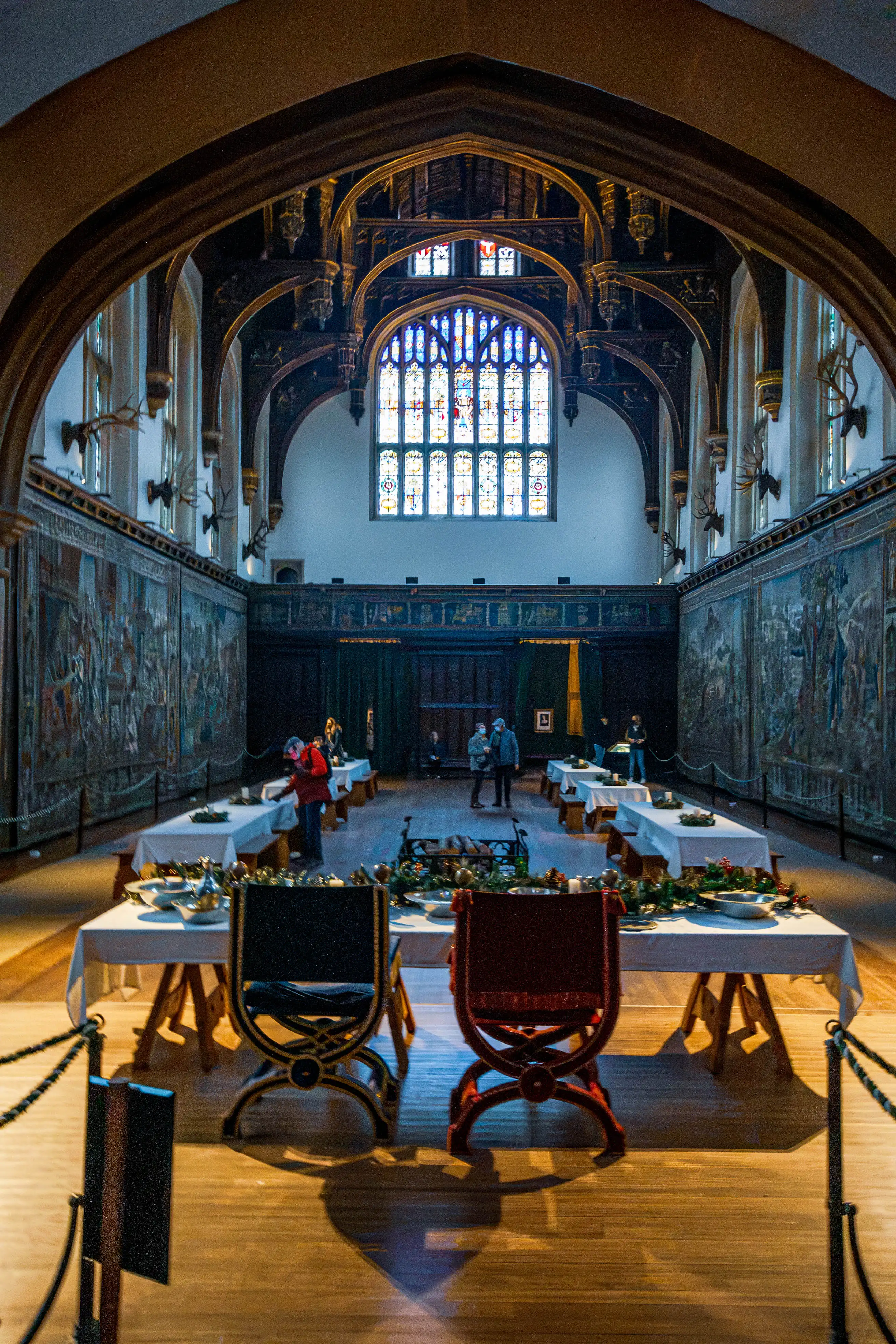 Great hall with vaulted timber ceiling, stained-glass window, tapestries, banquet tables and two ornate chairs with festive decoration.