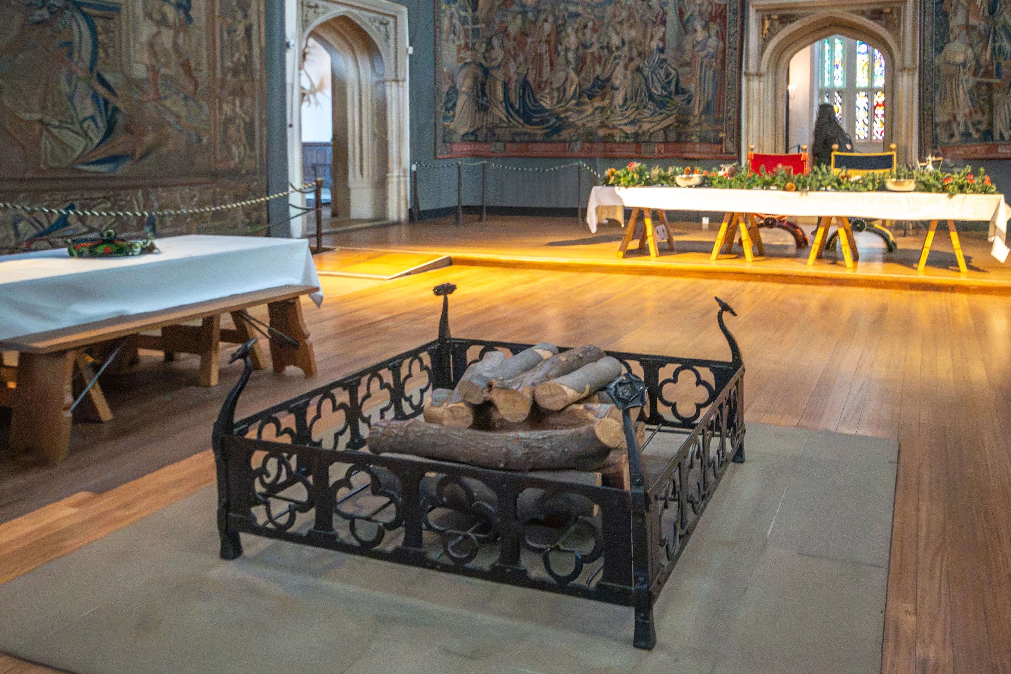 Great hall with wrought-iron brazier, banquet tables, greenery and fruit decoration, large tapestries, arched stone doorways and stained-glass windows.