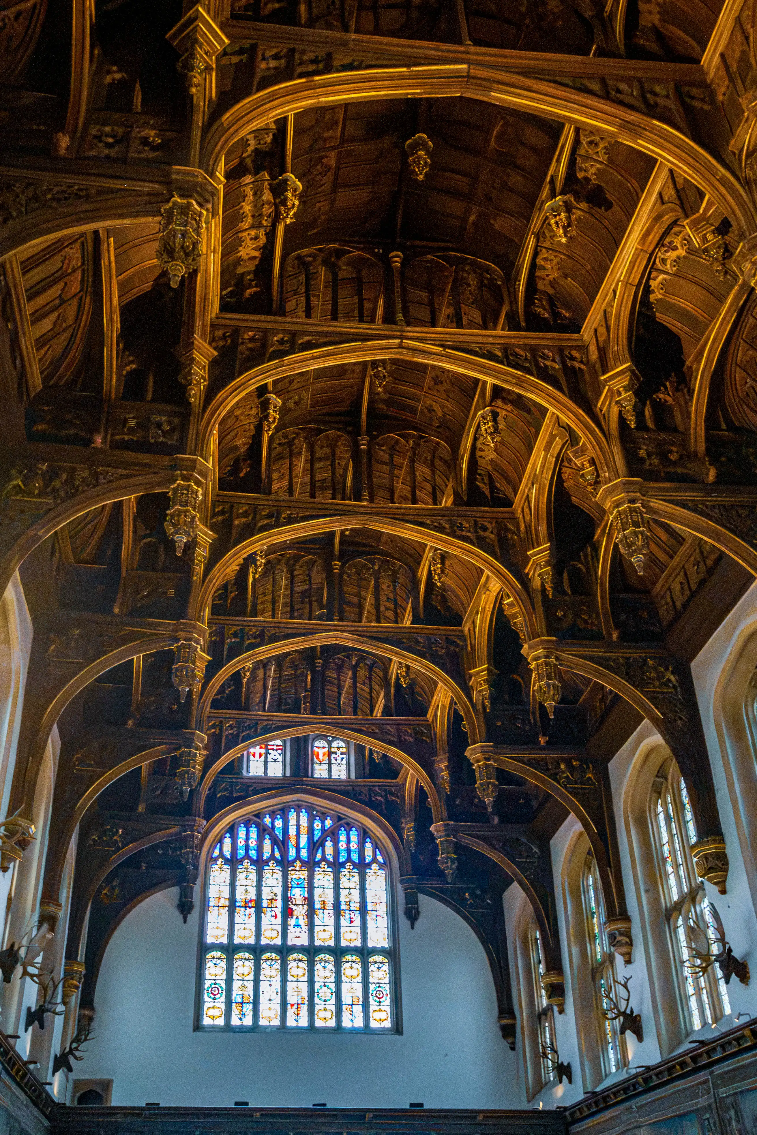 Historic hall with carved timber vaulting, gold highlights, stained-glass window, tall side windows and mounted antlers.