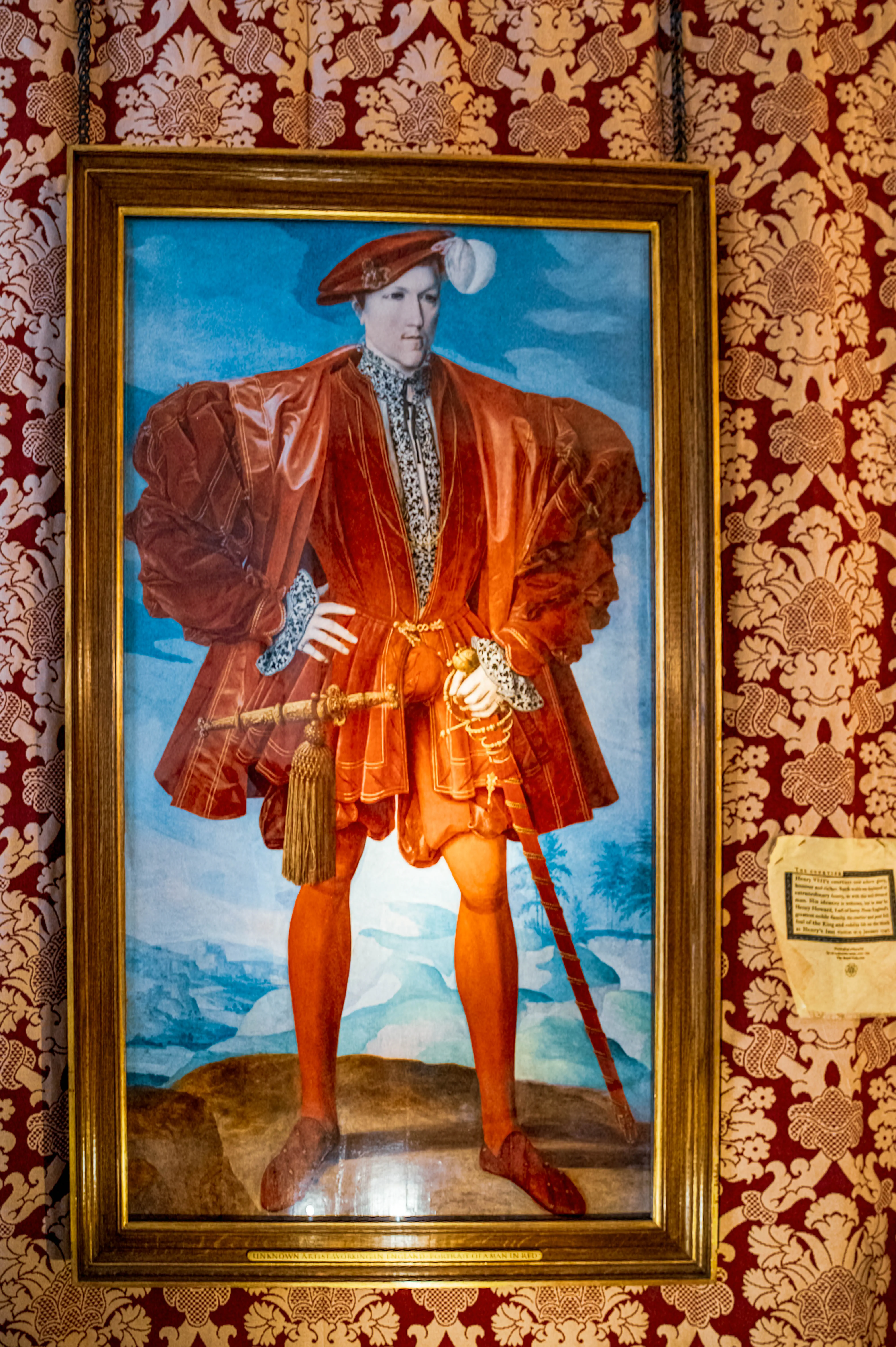 Renaissance portrait of a figure in red attire with feathered hat and sword, framed in gold on patterned wallpaper.