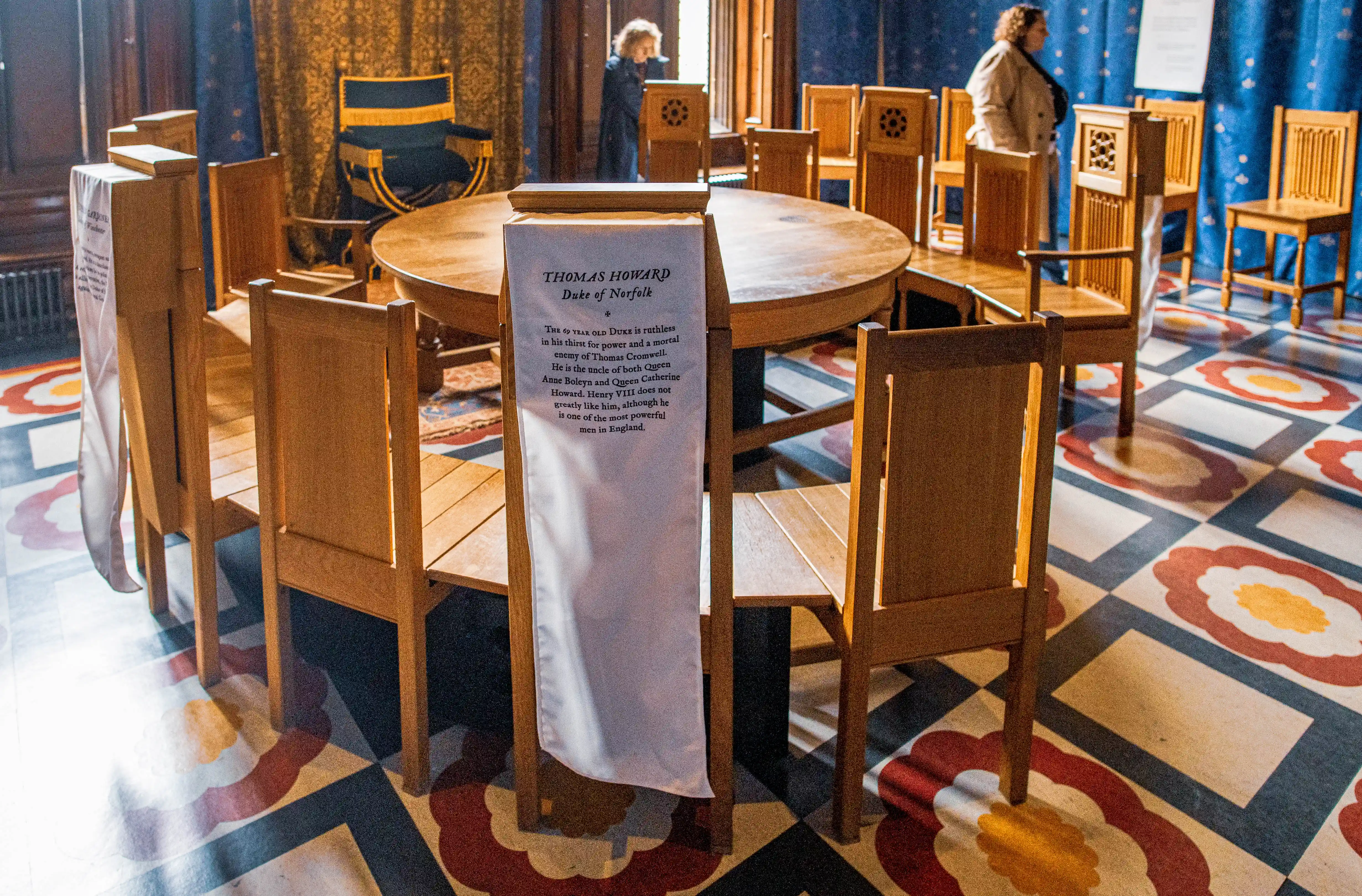 Historic room with round wooden table, chairs draped with interpretive banners, patterned floor and blue-and-gold curtains.