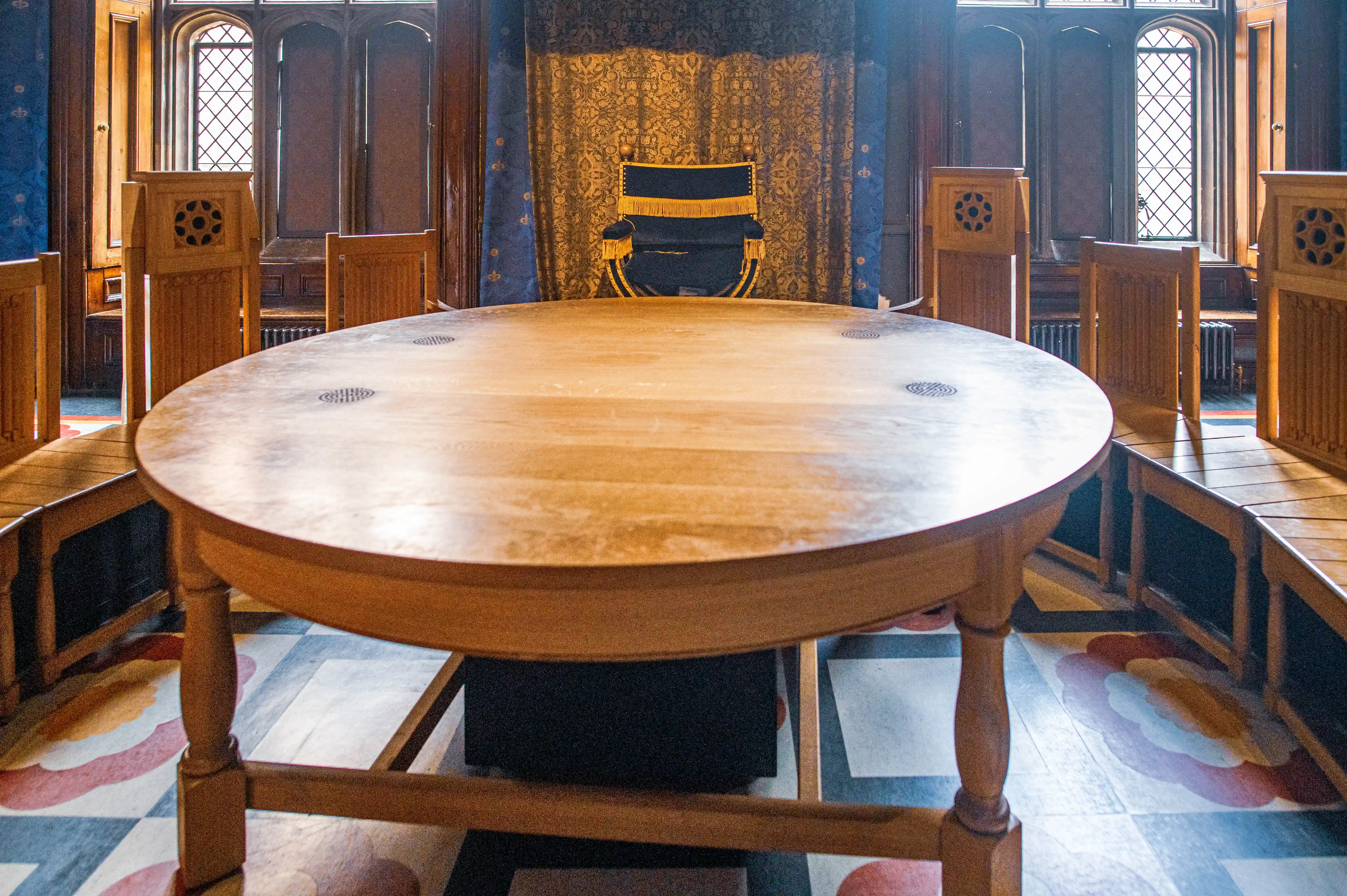 Historic chamber with round wooden table, decorative chairs, ornate blue-and-gold seat, patterned floor and arched windows.
