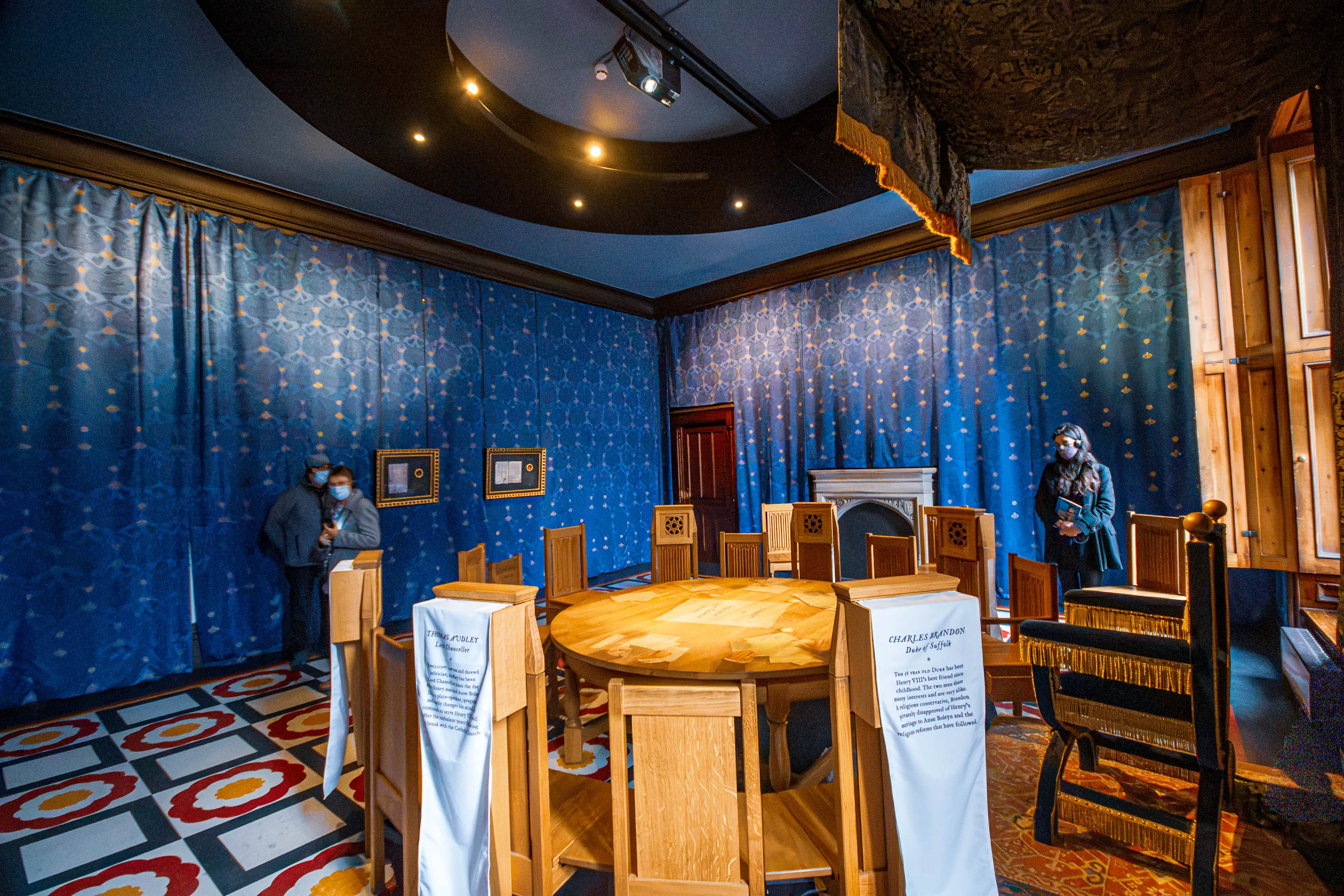 Historic chamber with round wooden table, chairs draped with interpretive banners, blue patterned walls, patterned floor and visitors wearing masks.