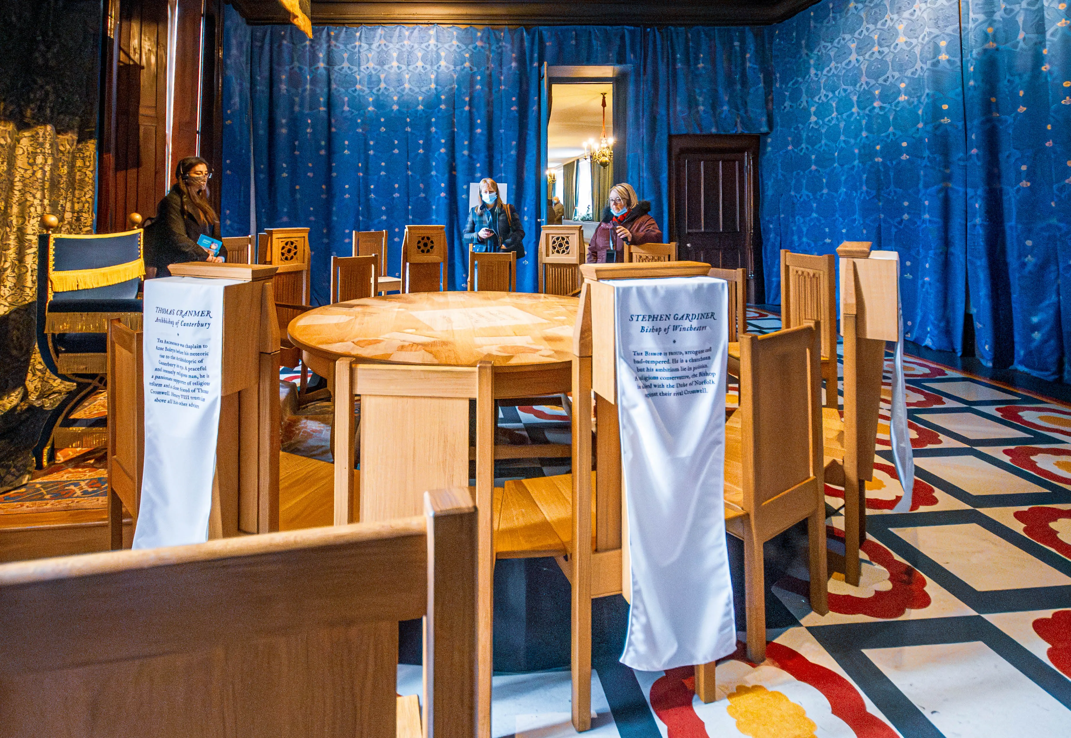 Historic chamber with round wooden table, chairs draped with interpretive banners, blue patterned walls, patterned floor and visitors wearing masks.