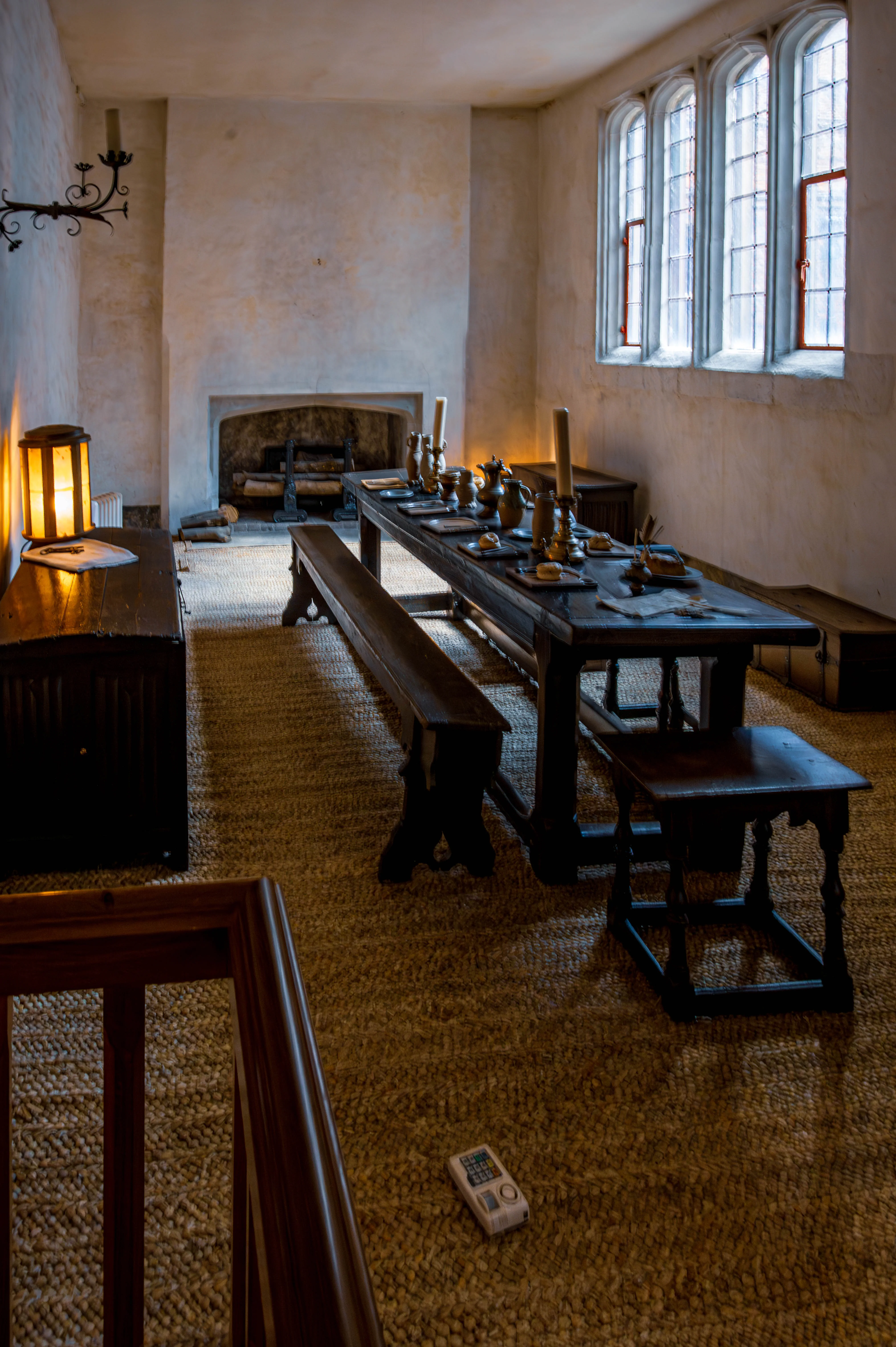 Historic dining hall with long wooden table, pewter tableware, stone fireplace, tall windows and woven-mat floor.