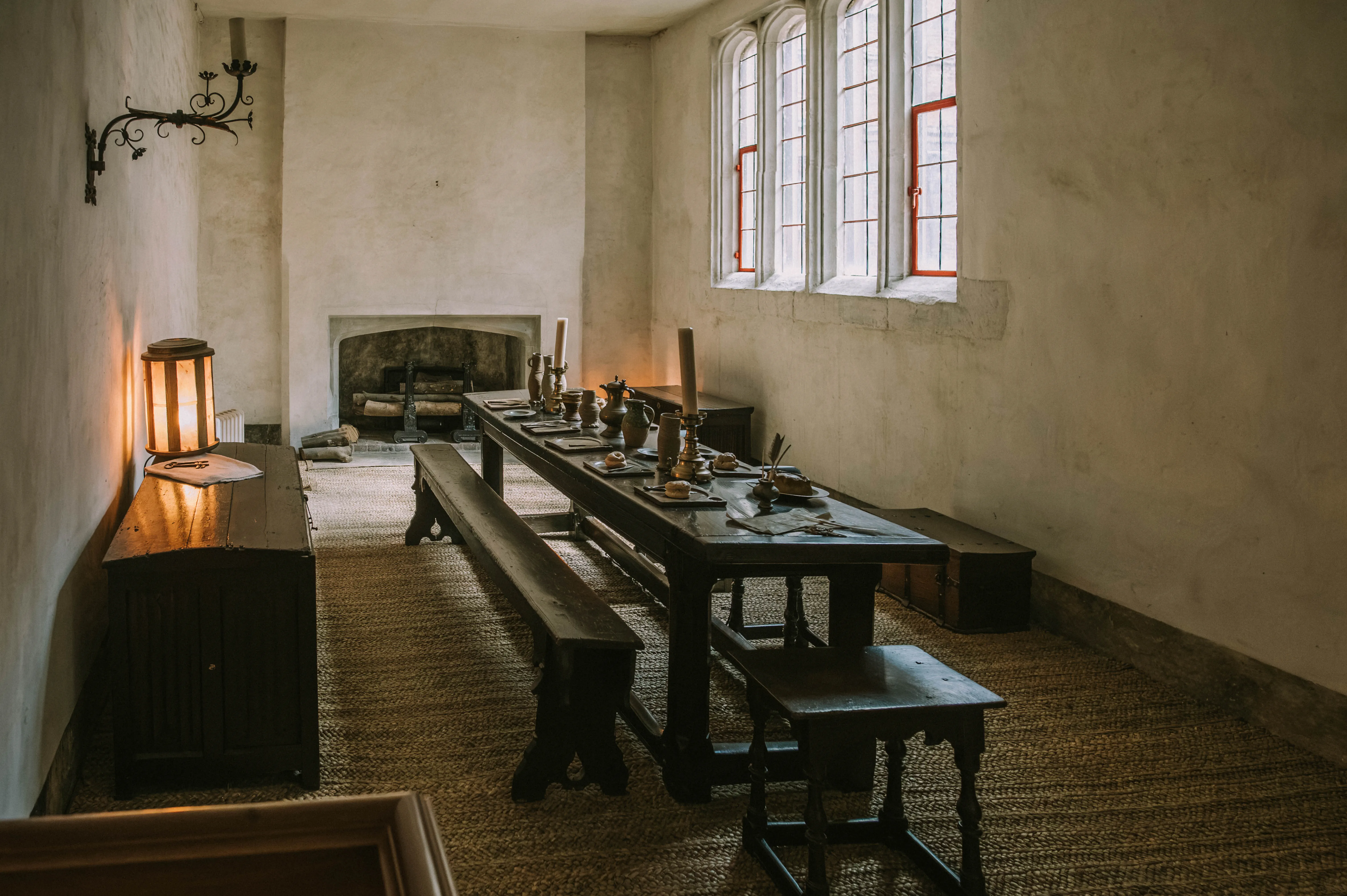 Historic dining room with long wooden table, pewter tableware, stone fireplace, tall-paned window and woven-mat floor.