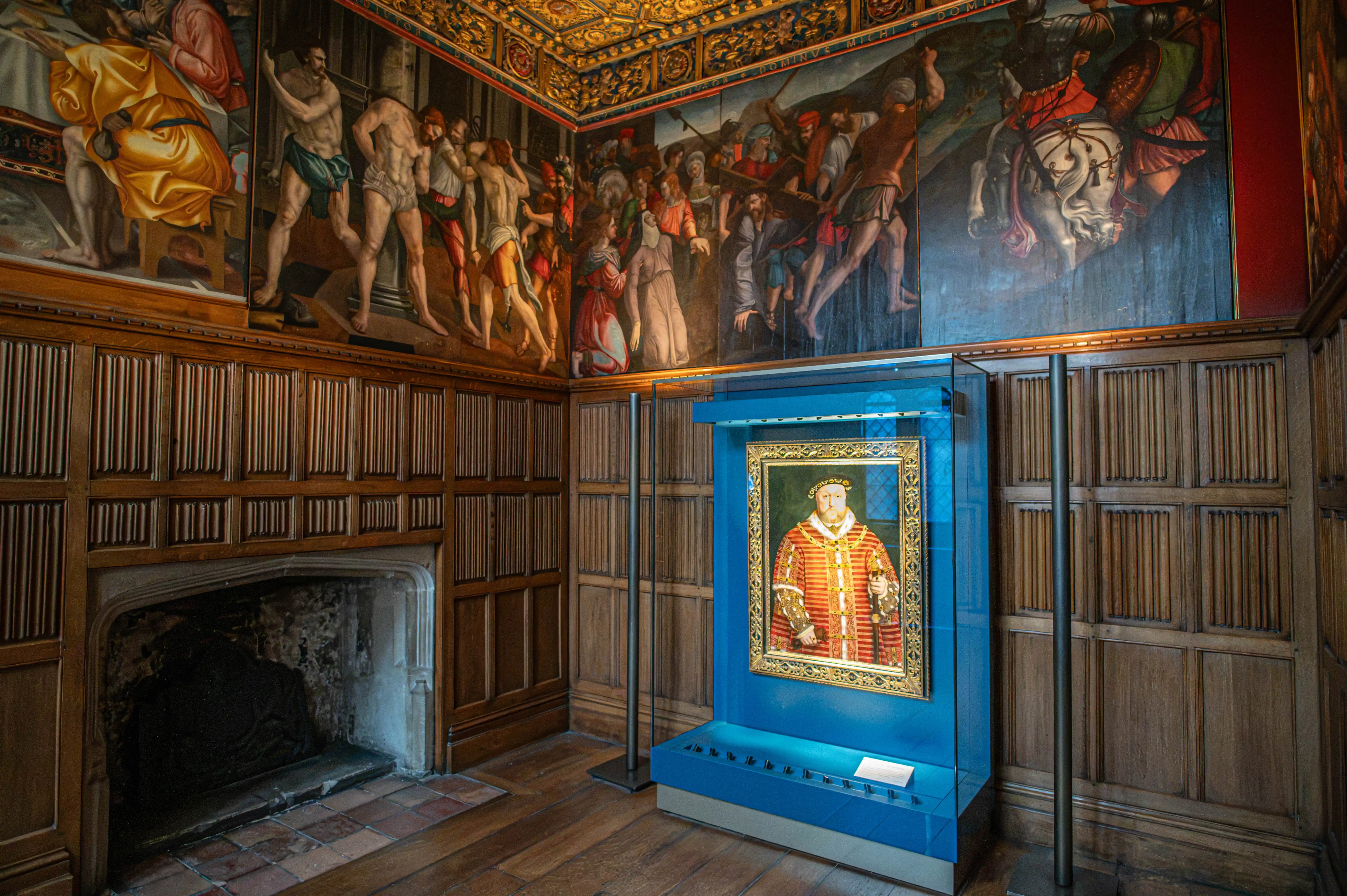 Historic room with Renaissance-style paintings, carved panelling, gold ceiling, stone fireplace and framed regal portrait in a blue display case.