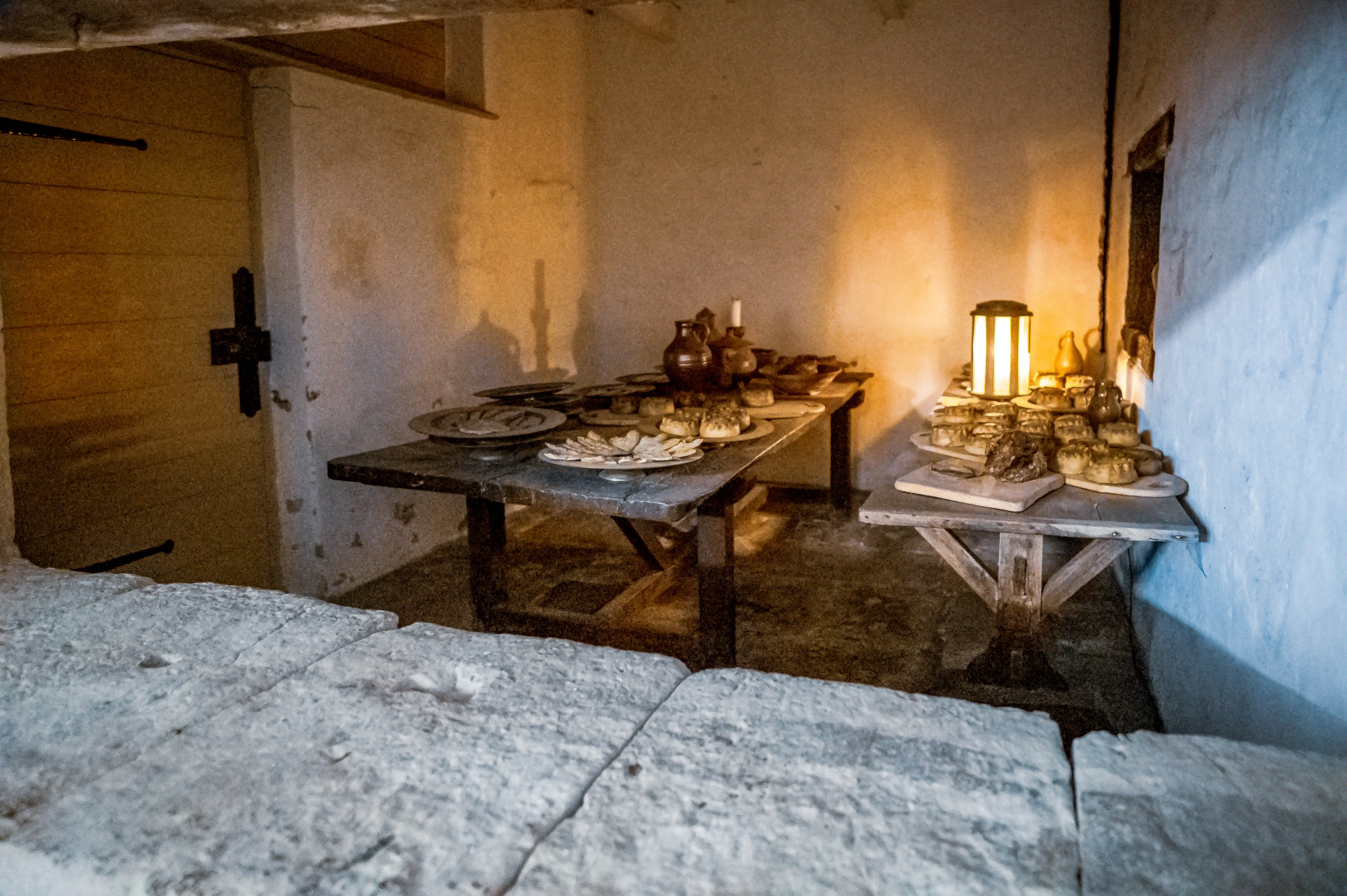 Rustic kitchen with wooden tables, pies, pastries, ceramic jugs and warm lantern light.