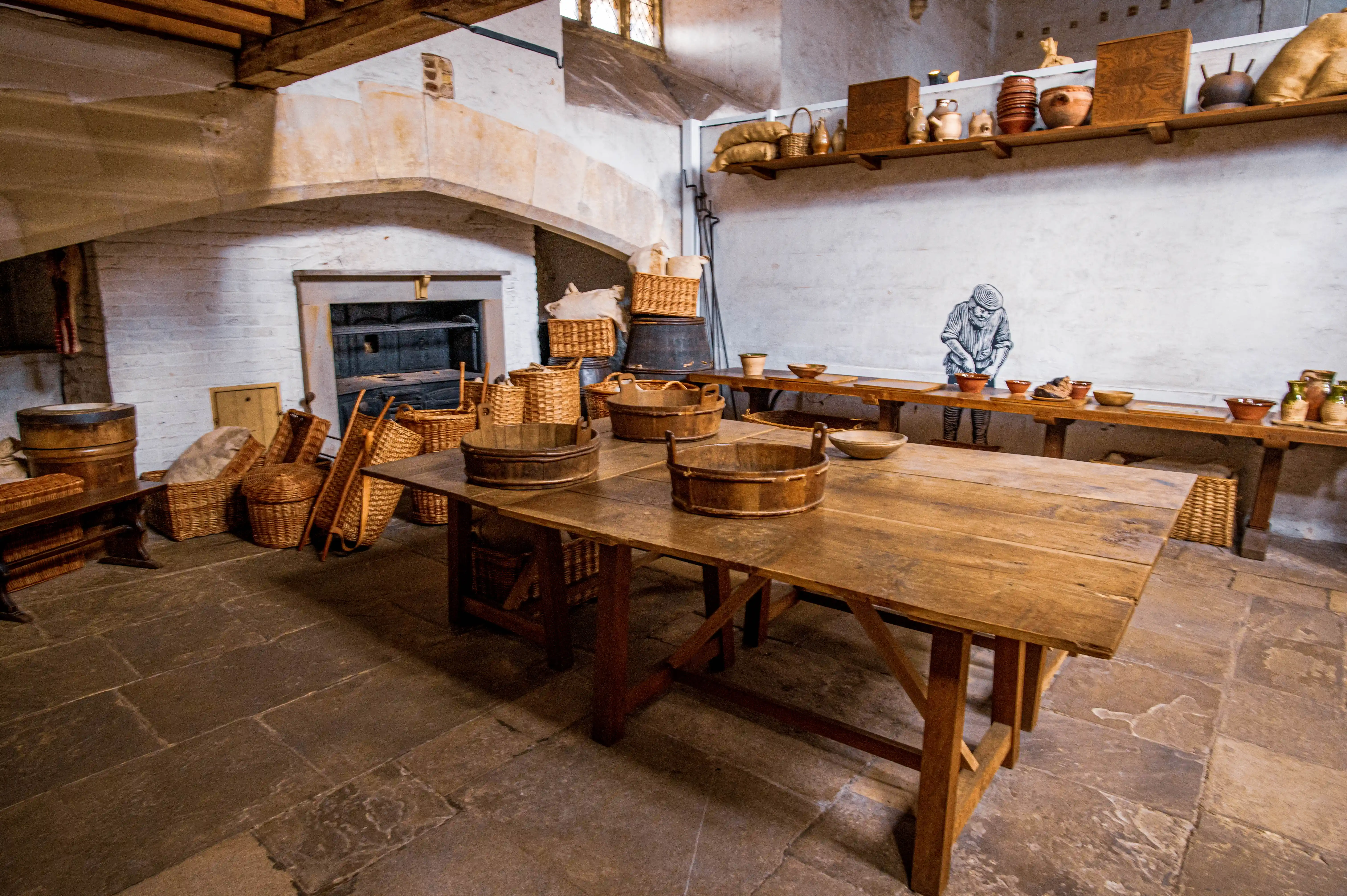 Historic kitchen with stone arch, wooden table, tubs, baskets, barrels and pottery on shelves