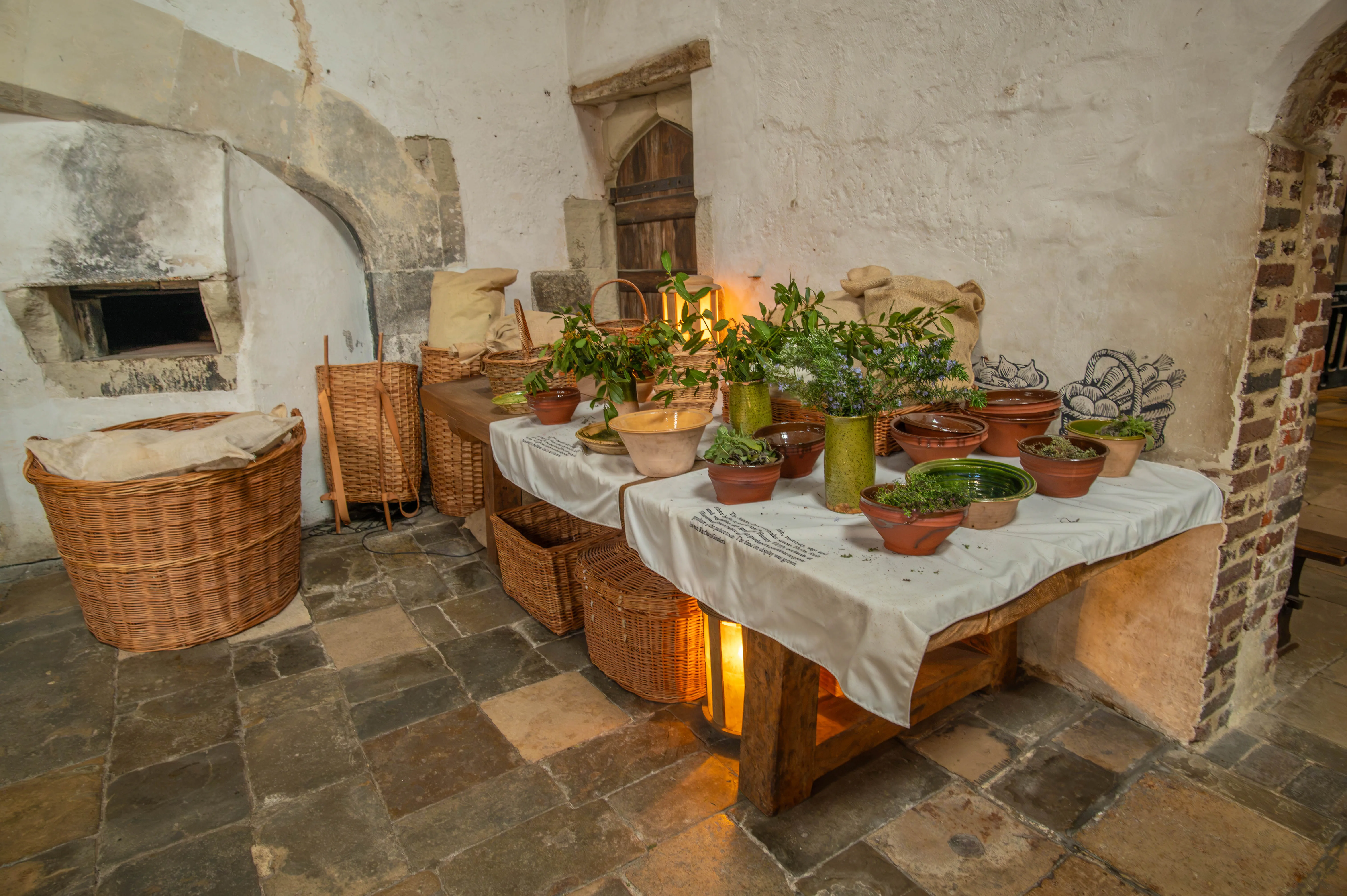 Rustic historic kitchen with pottery vessels, herbs, wicker baskets, cloth sacks and stone hearth.