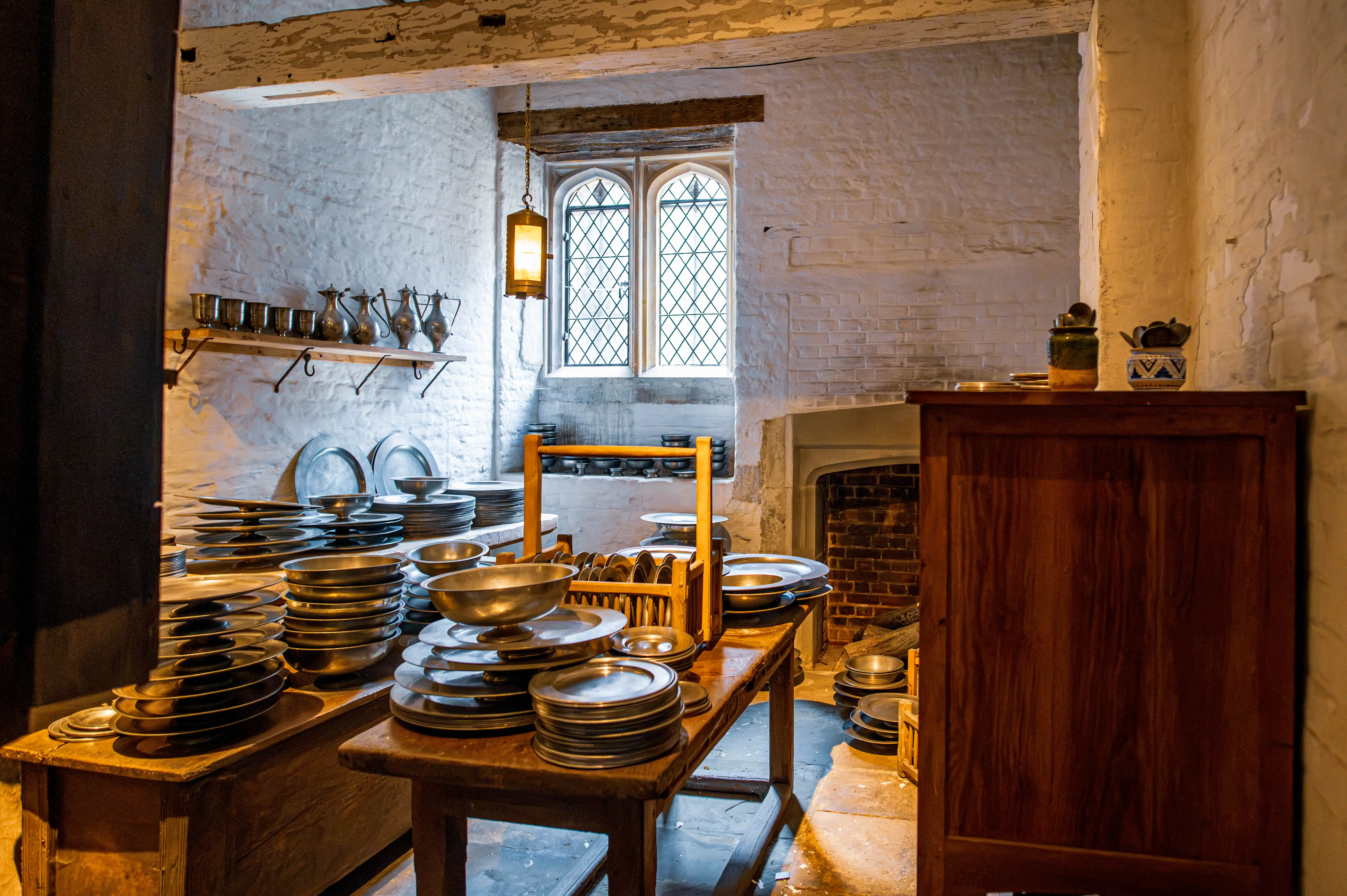 Historic scullery with pewter plates, wooden tables, shelves of pitchers, cabinet, diamond-paned window and lantern.