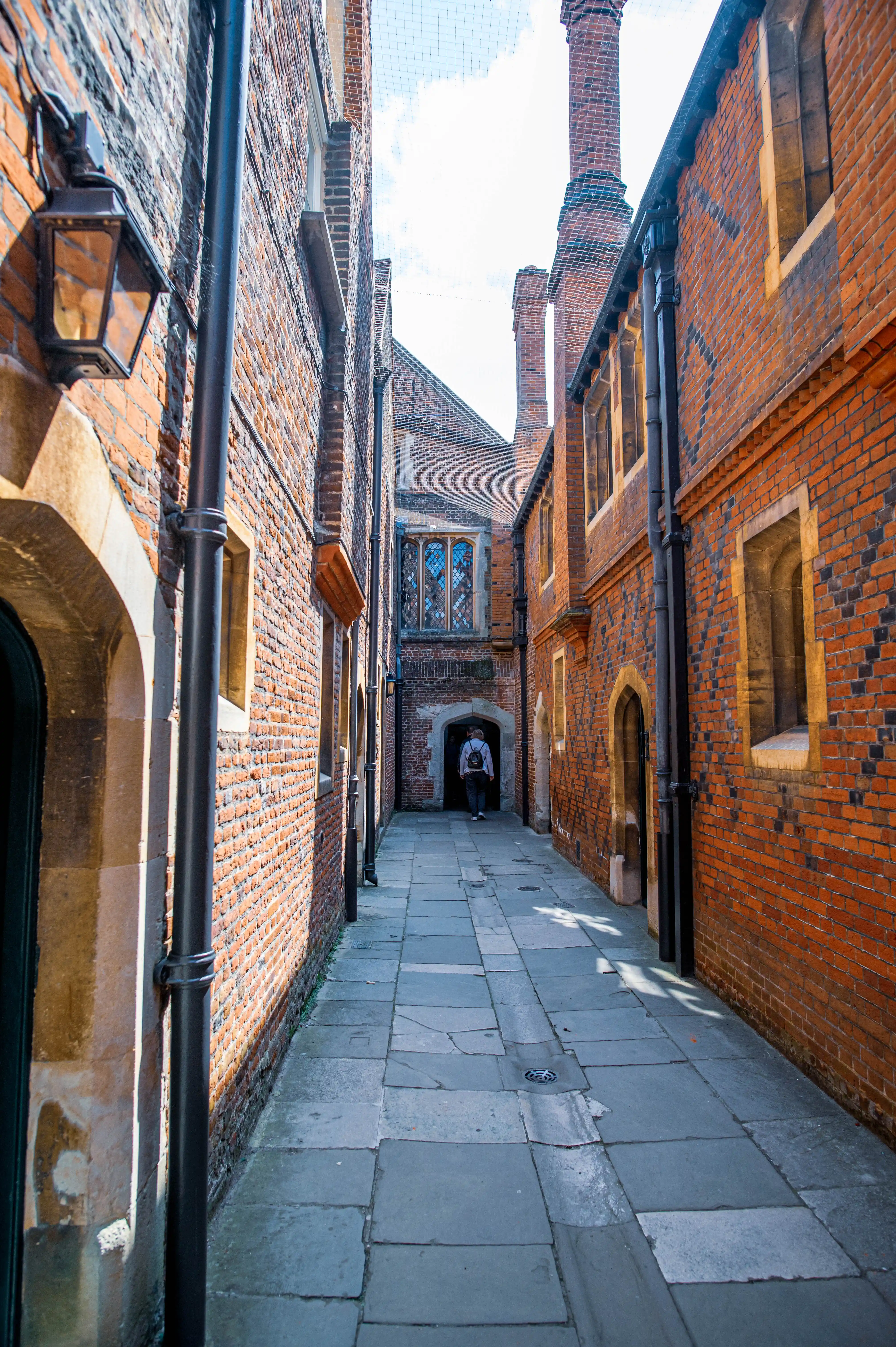 A narrow alleyway between tall historic brick buildings featuring stone-framed windows, arched doorways and prominent chimneys. Stone-slab paving leads toward an archway at the far end, where a person walks away. Drainpipes and lantern-style wall lights add detail to the traditional architectural setting, illuminated by daylight with soft shadows across the ground.