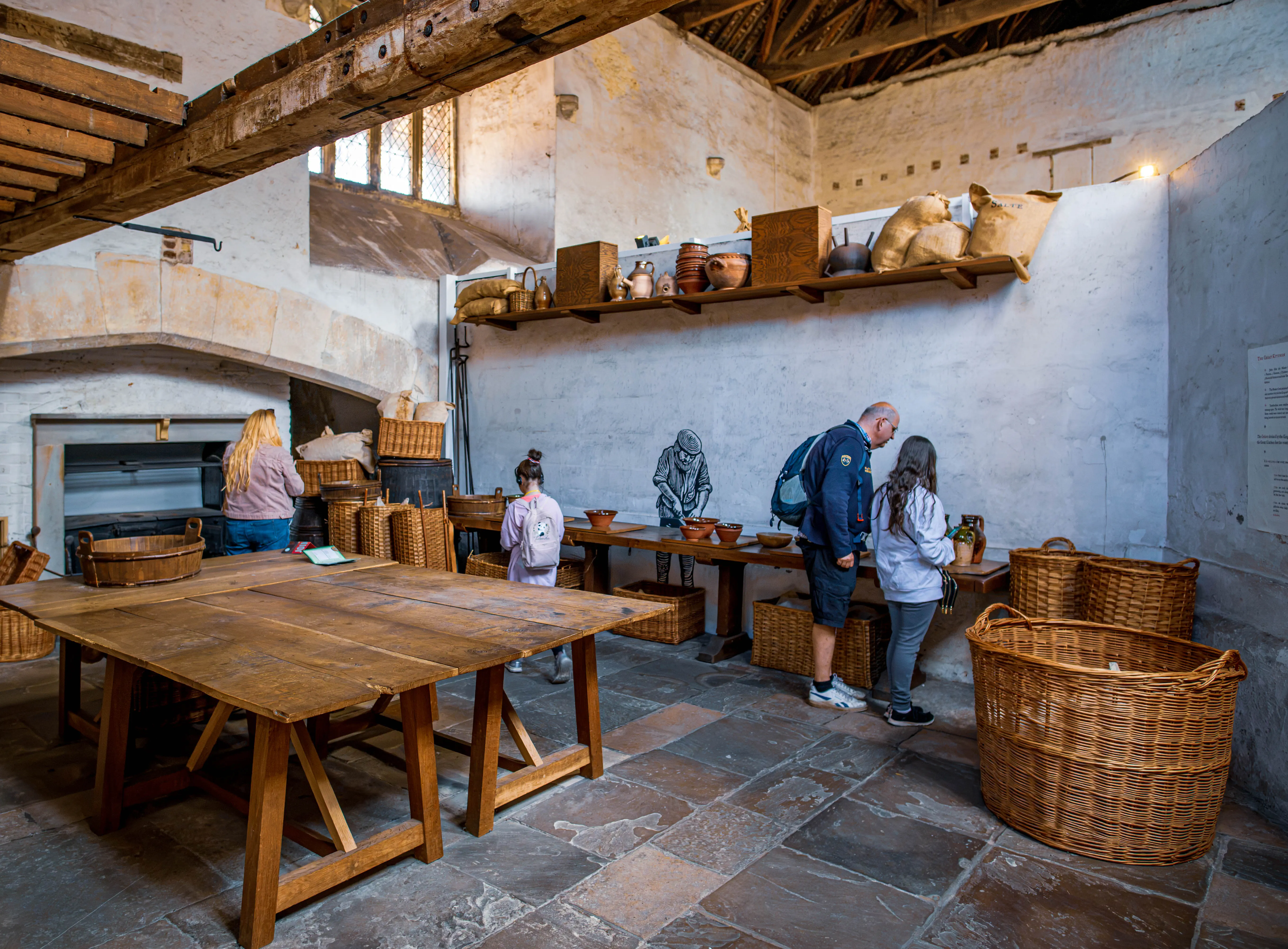 Historic kitchen storeroom with wicker baskets, wooden tables, pottery on shelves and visitors observing the display.