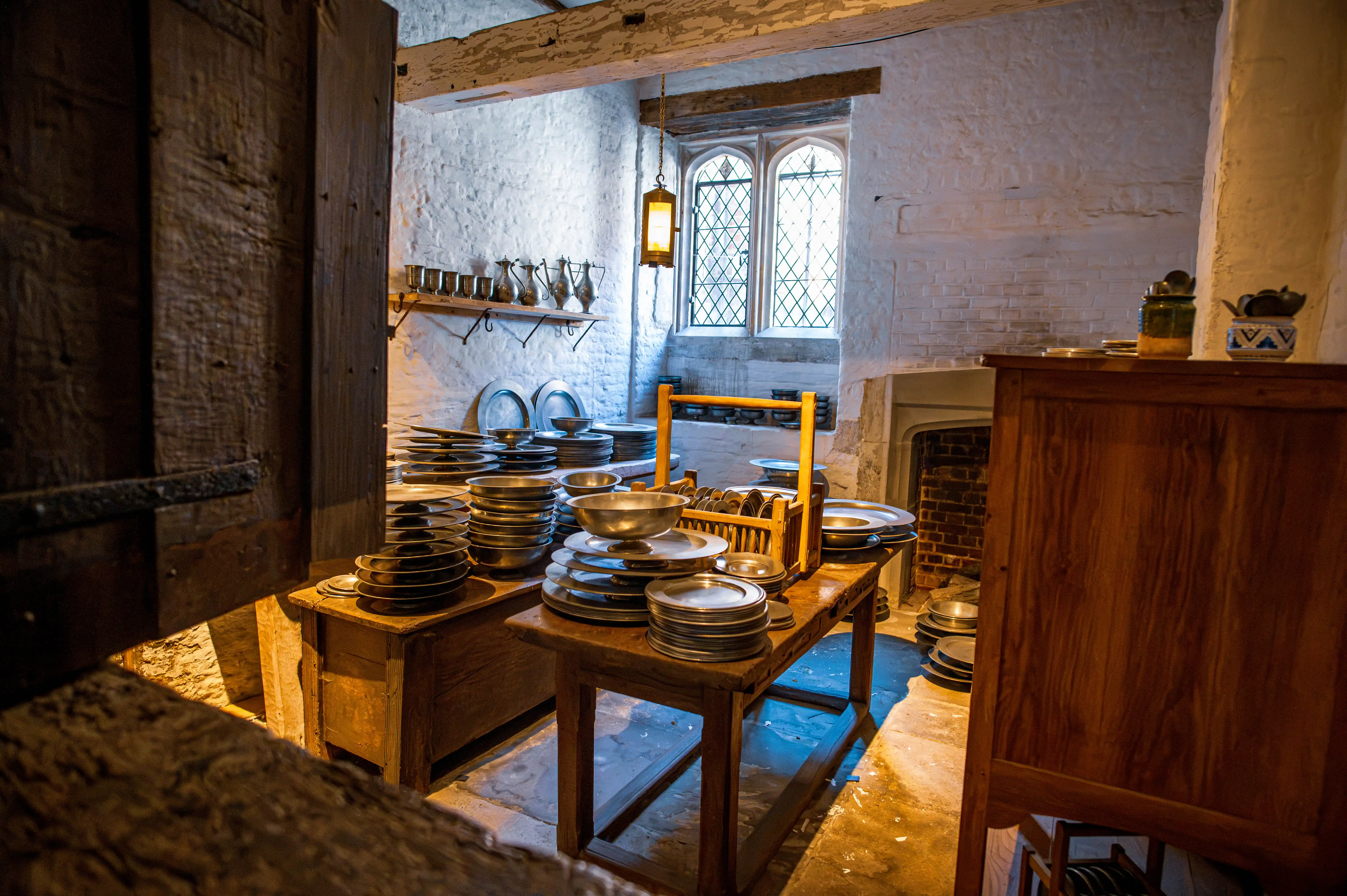 Historic scullery with pewter plates, wooden tables, shelves of pitchers, diamond-paned window, lantern and hearth.