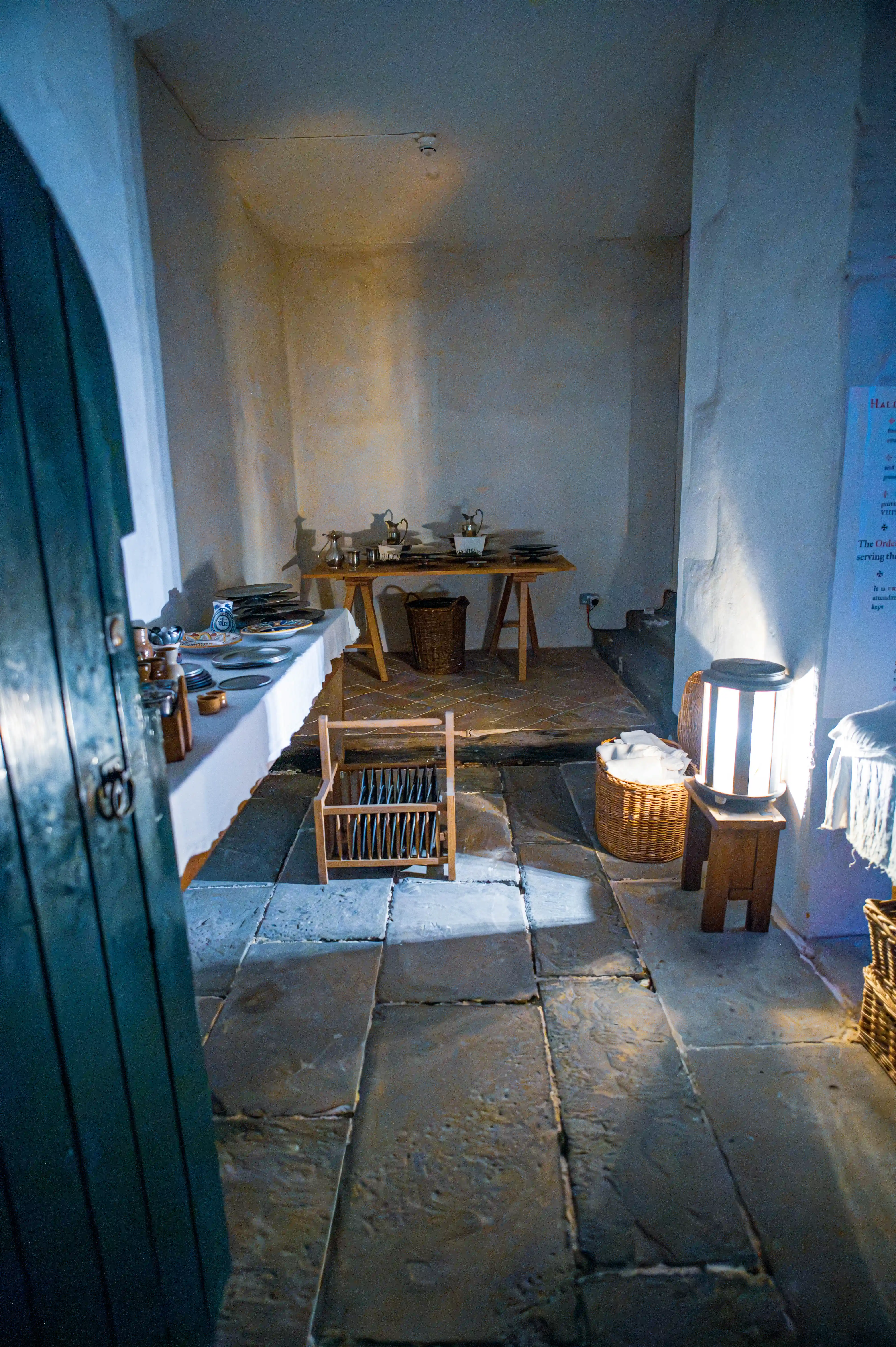 Historic kitchen interior with stone floor, wooden tables, wicker baskets, plate rack and lantern light.
