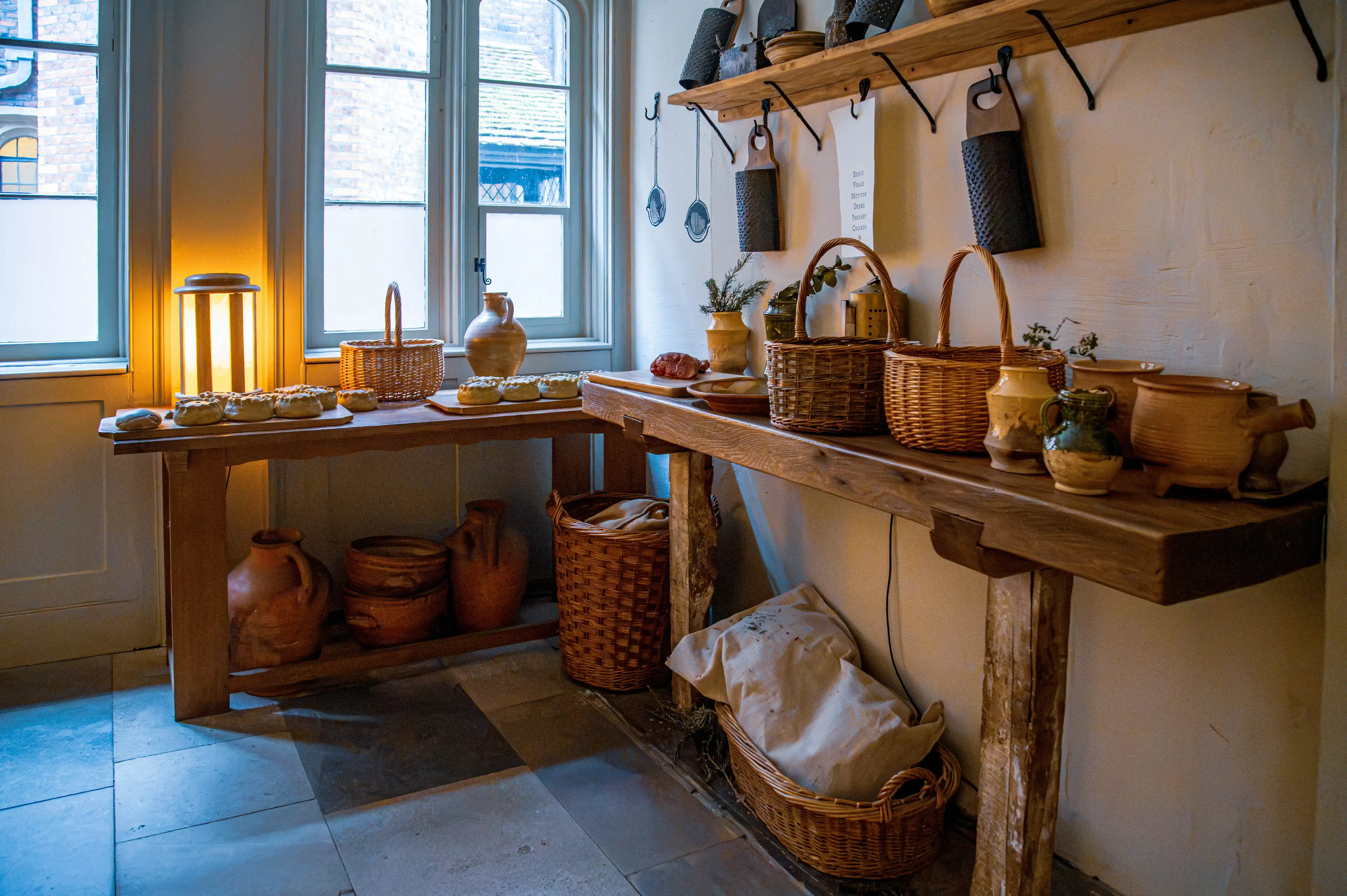 Rustic pantry with wooden tables, woven baskets, ceramic pots, herbs, metal utensils and warm lamp light.