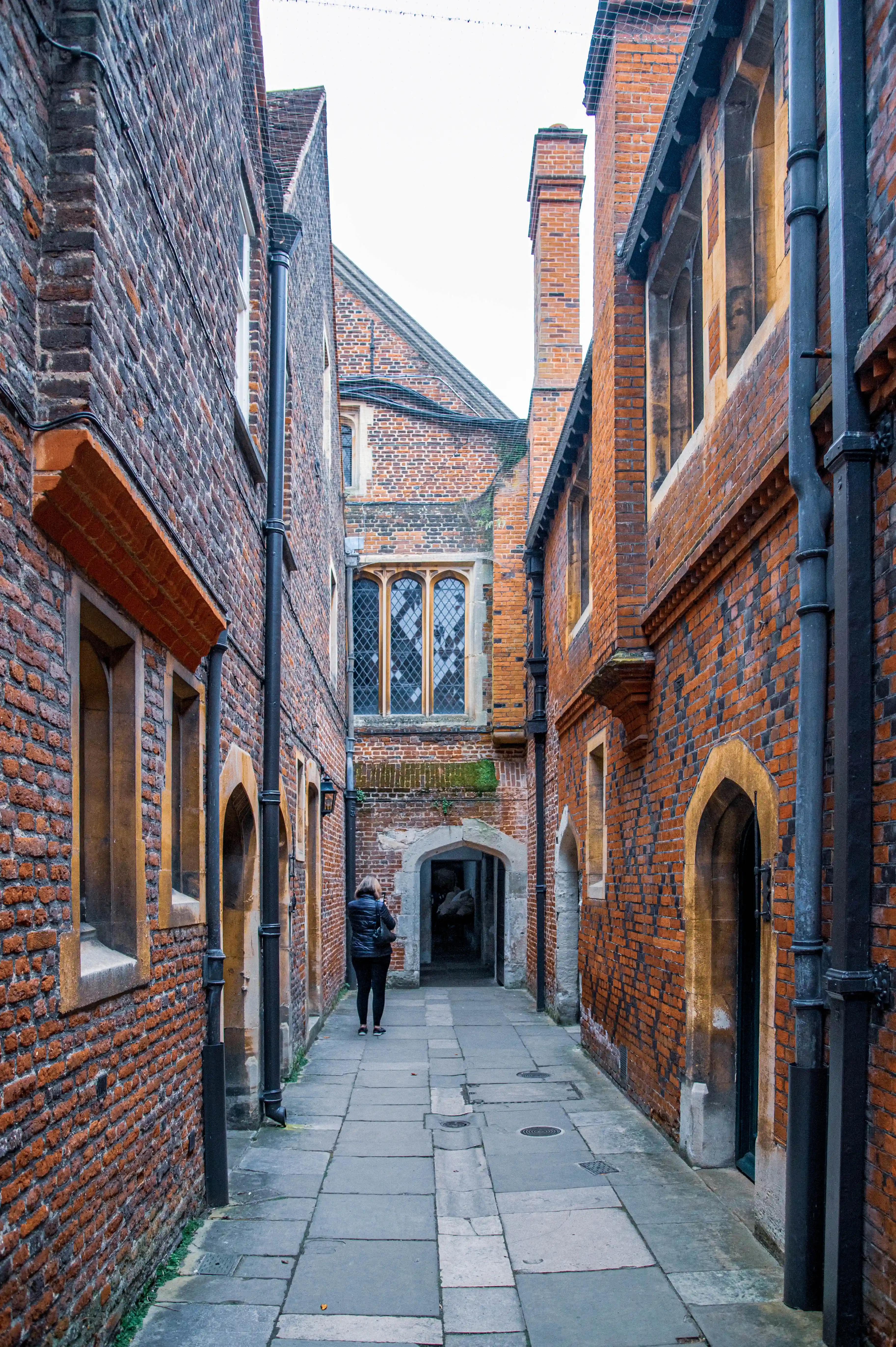 Narrow historic brick alleyway with stone-framed leaded windows, arched doorway and a person walking away