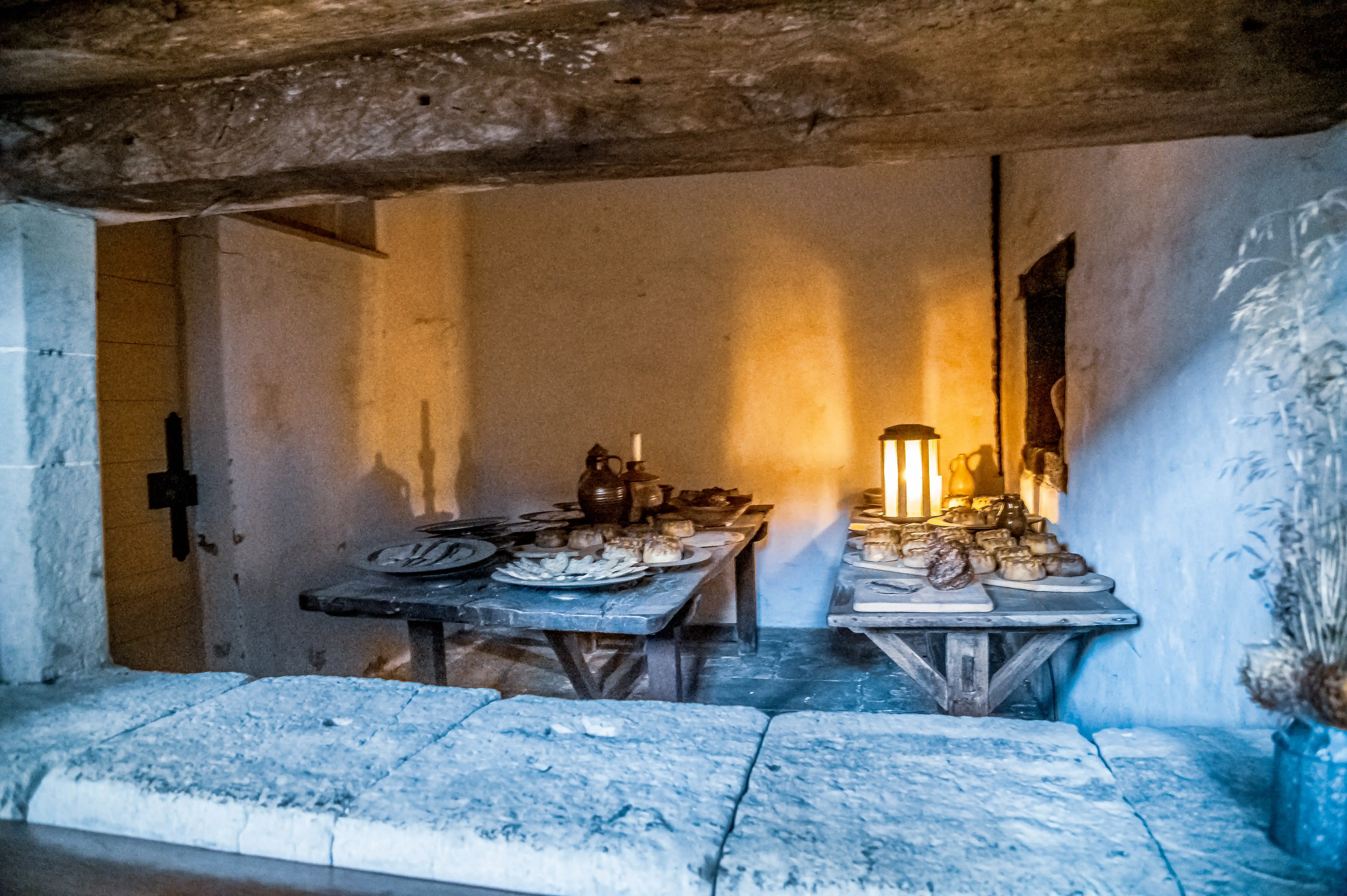 Rustic kitchen with wooden tables, bread, pies, ceramic vessels and candle lantern against stone walls.
