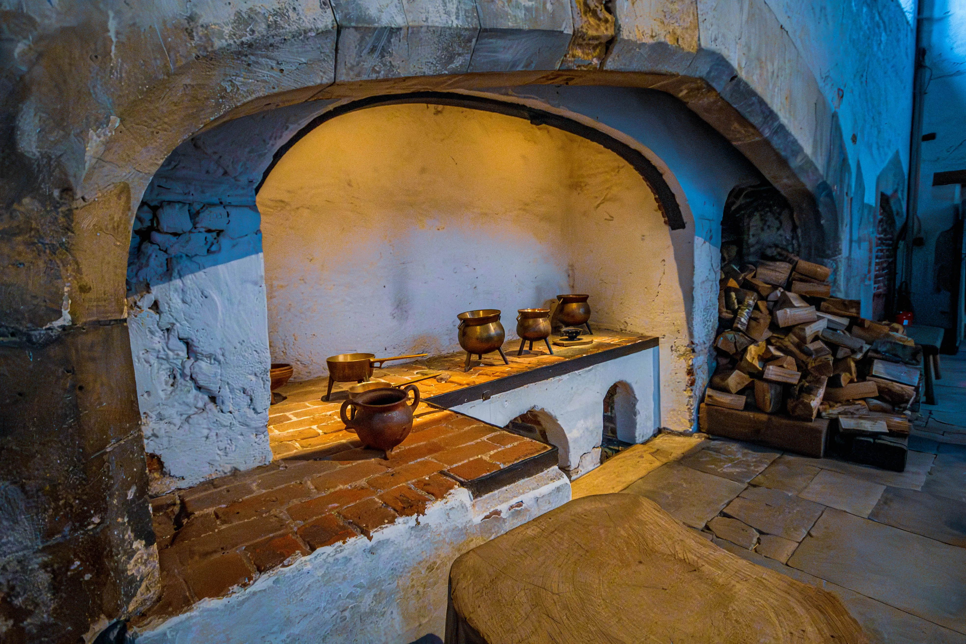 Historic cooking hearth with bronze pots on brick stands, stacked firewood and arched stone opening.
