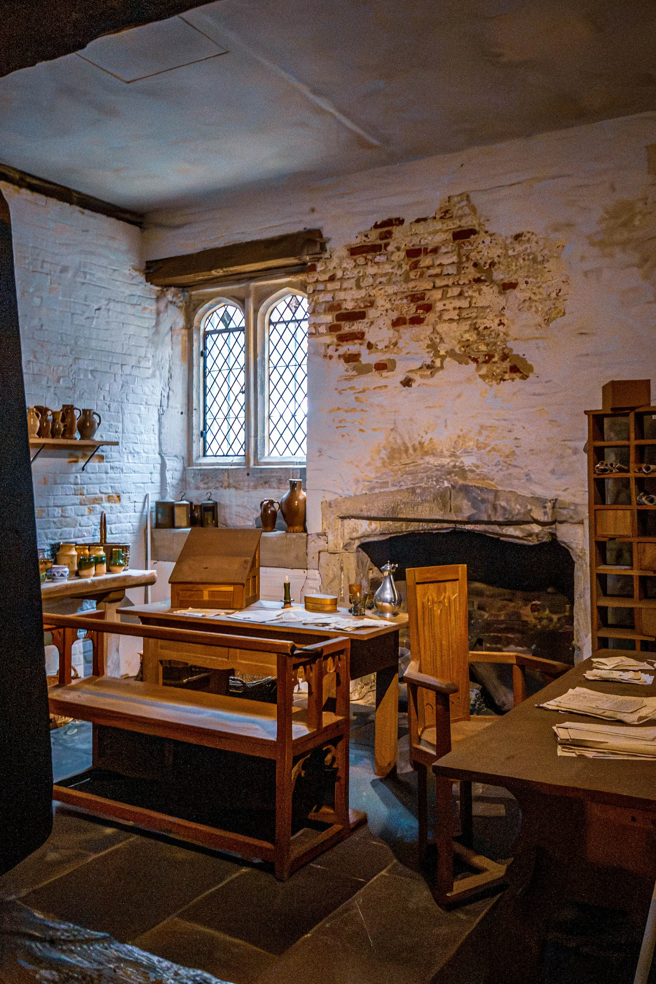 Historic study with writing table, pottery, glass bottles, stone fireplace and leaded-glass window.
