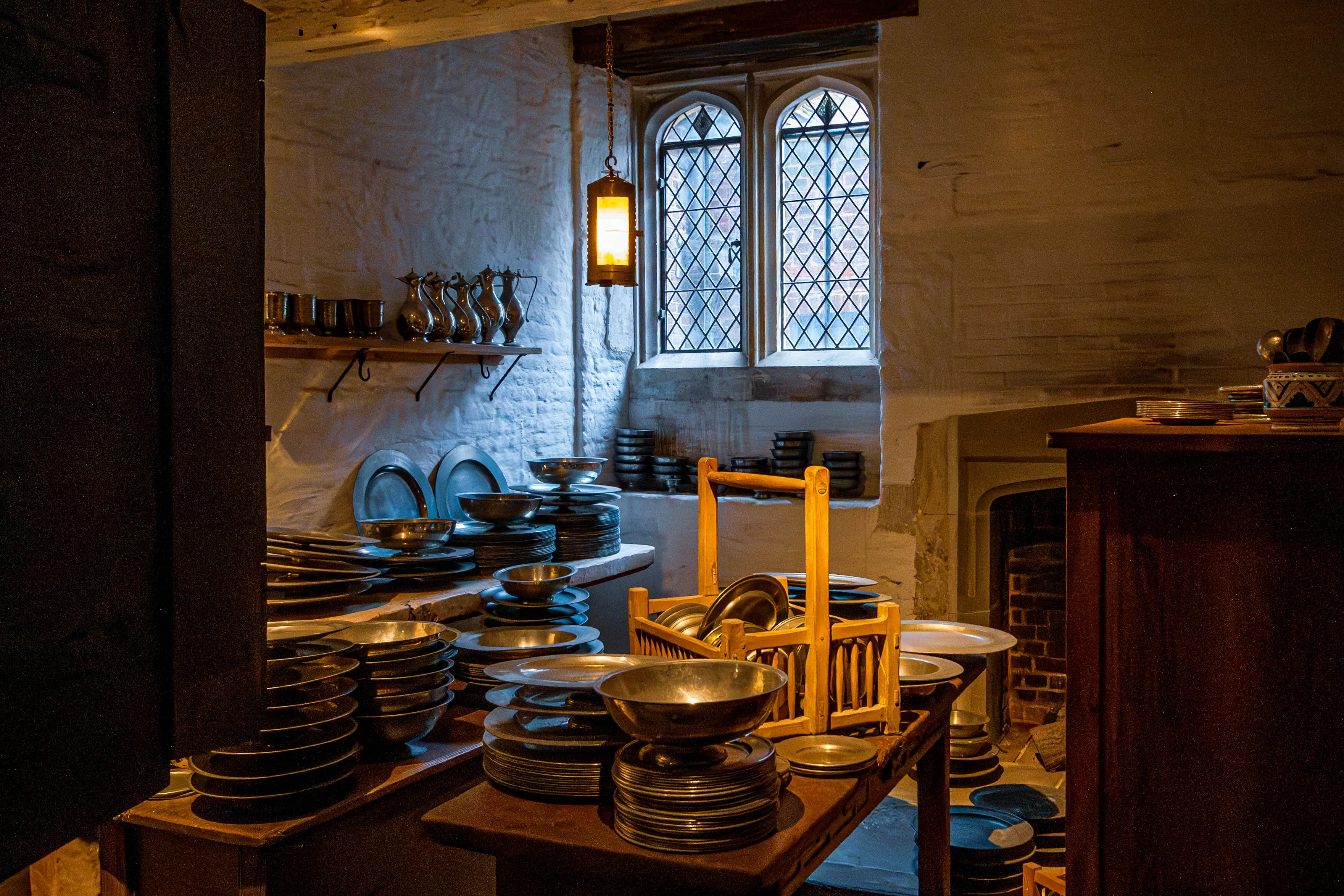 Historic scullery with pewter dishware, wooden beams, leaded-glass window and hanging lantern.
