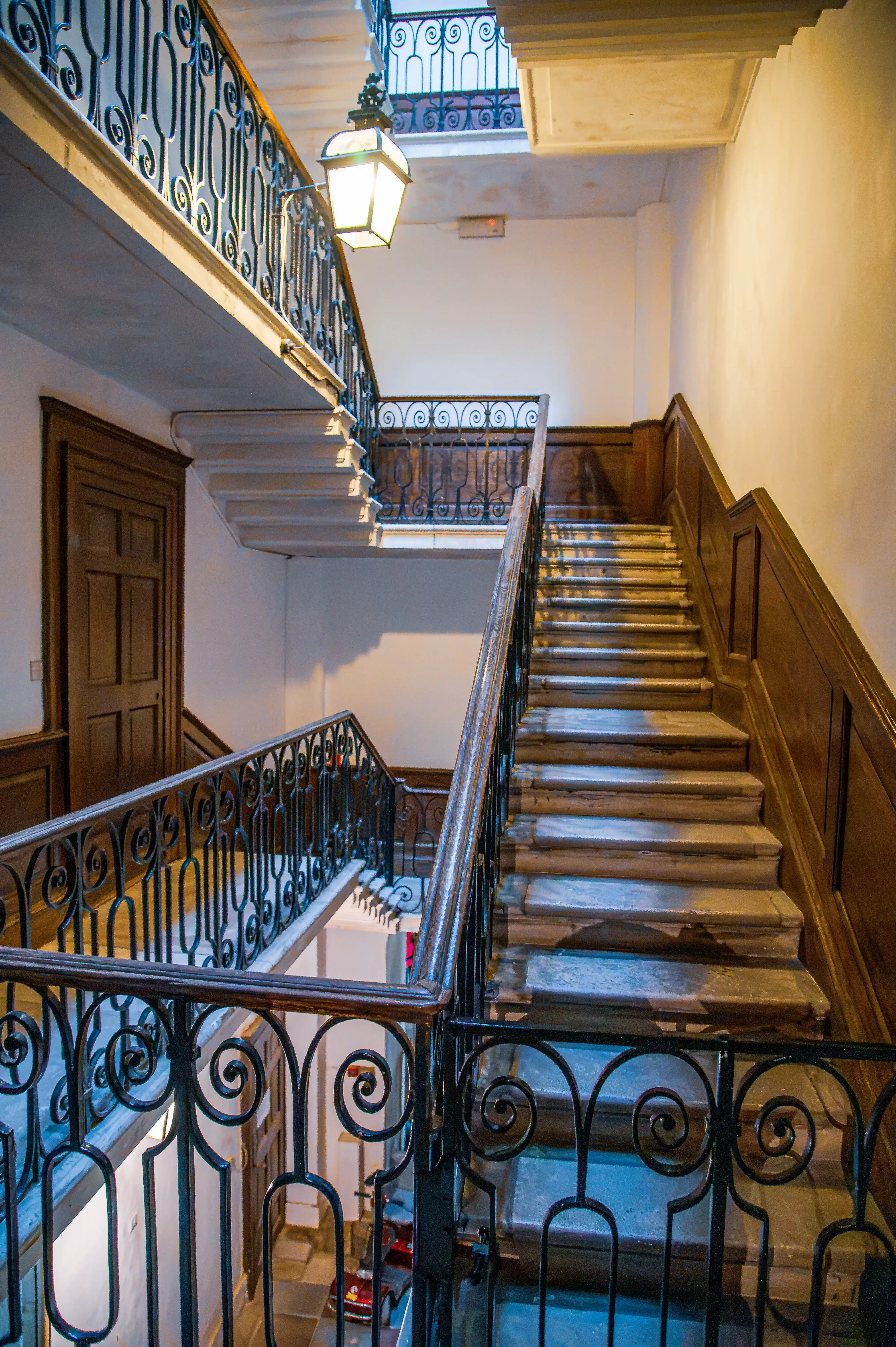 Staircase with ornate iron railings leading to a wooden door framed by a classical pediment and lit by a skylight.