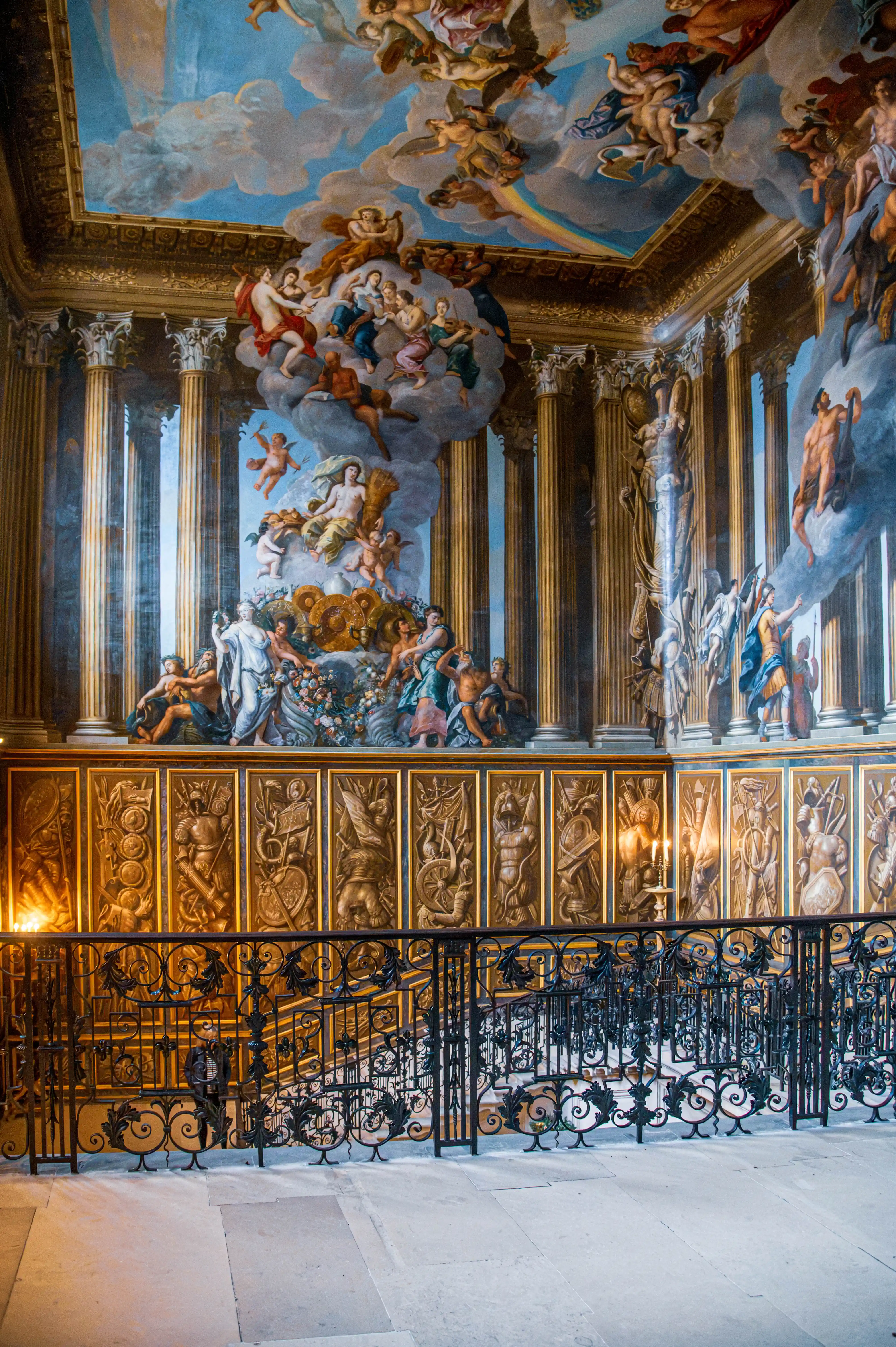 Grand staircase with ornate wrought-iron railings, gilded sculpted wall panels, classical columns and Baroque ceiling fresco.