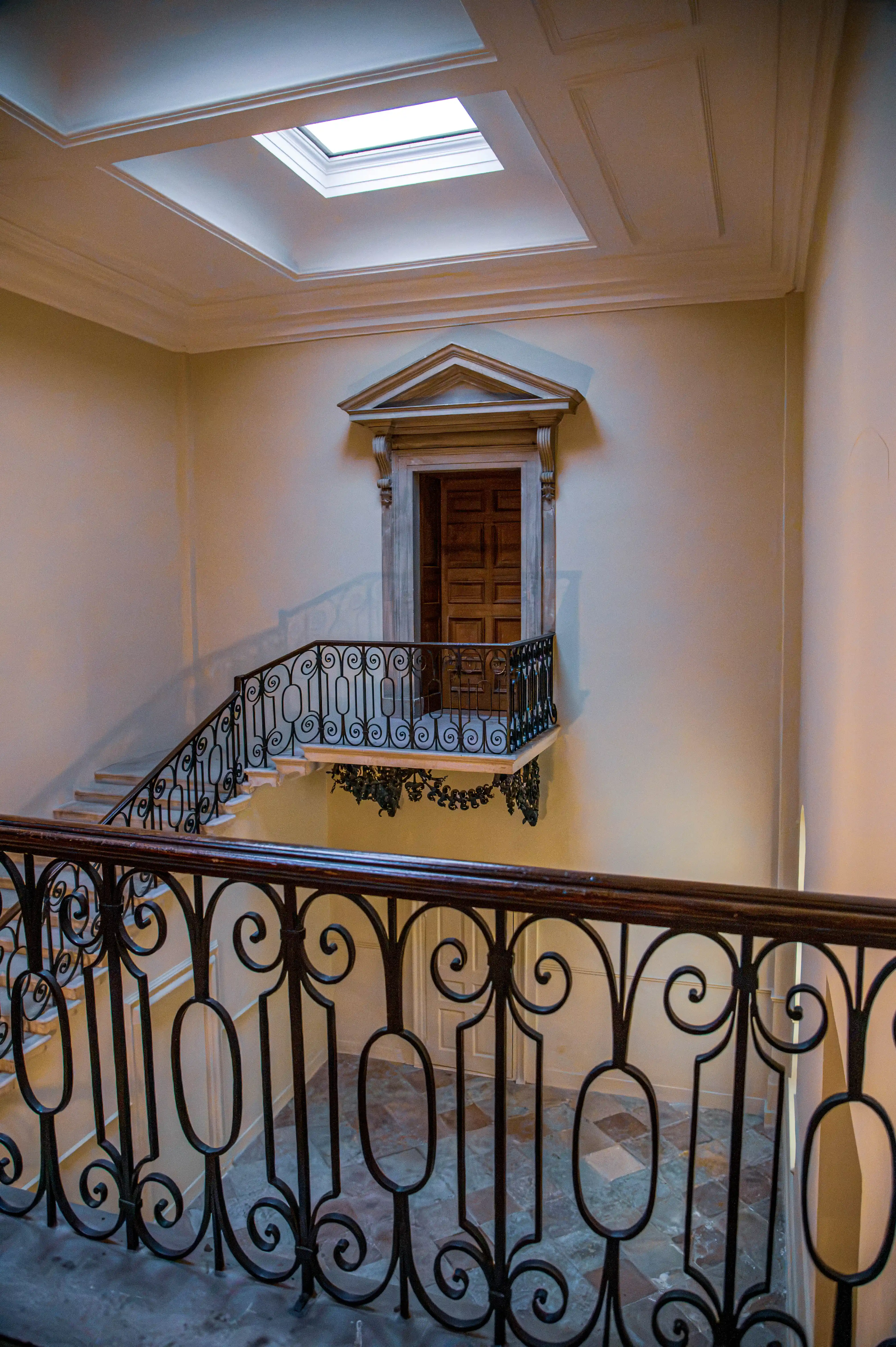 Staircase with ornate iron railings leading to a wooden door framed by a classical pediment and lit by a skylight.