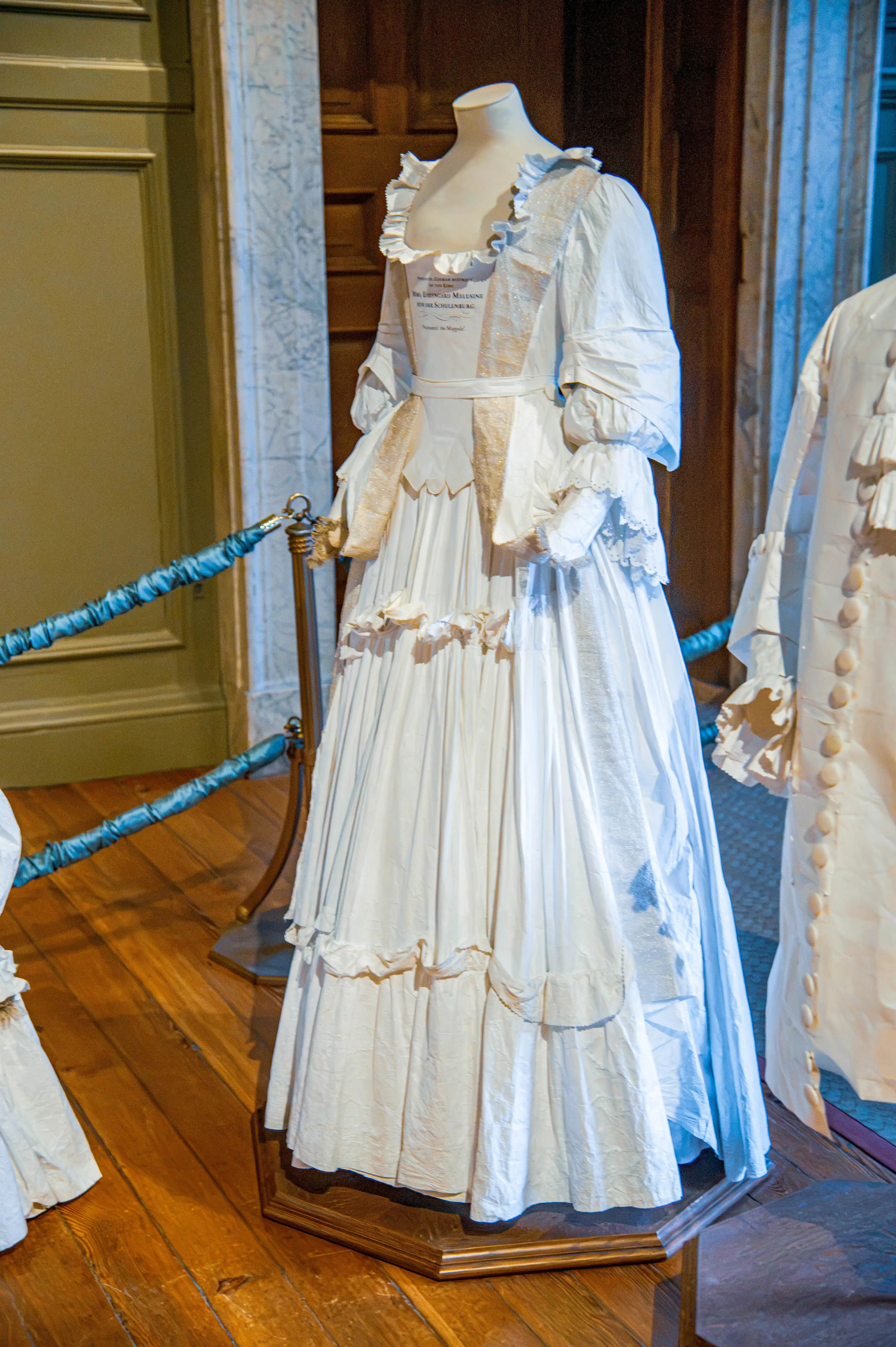 White historic gown with square neckline, puffed sleeves and tiered ruffled skirt displayed on a mannequin.
