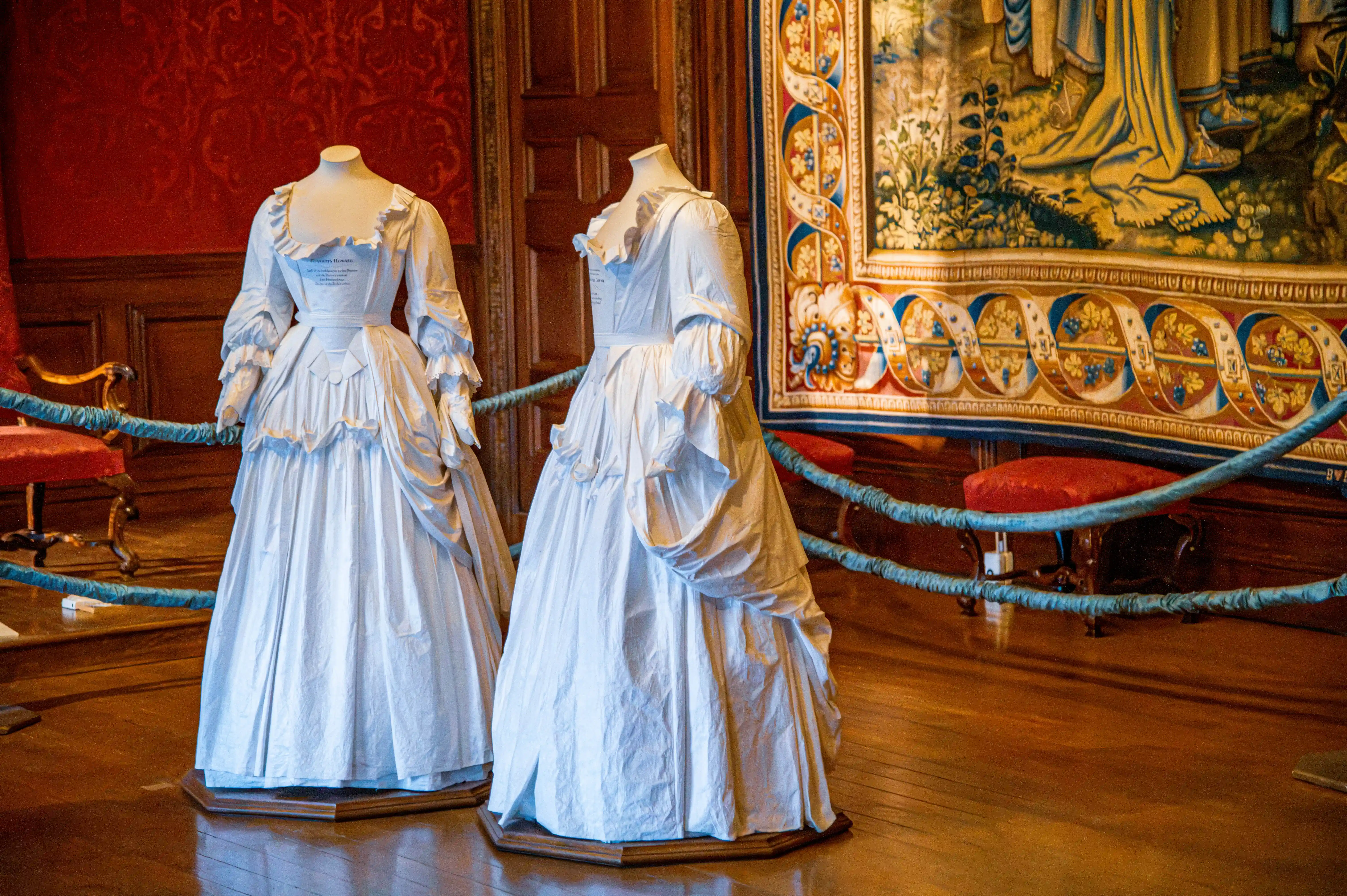Two light-coloured historic gowns on mannequins with ruffled detailing, displayed in front of a tapestry and antique furniture.