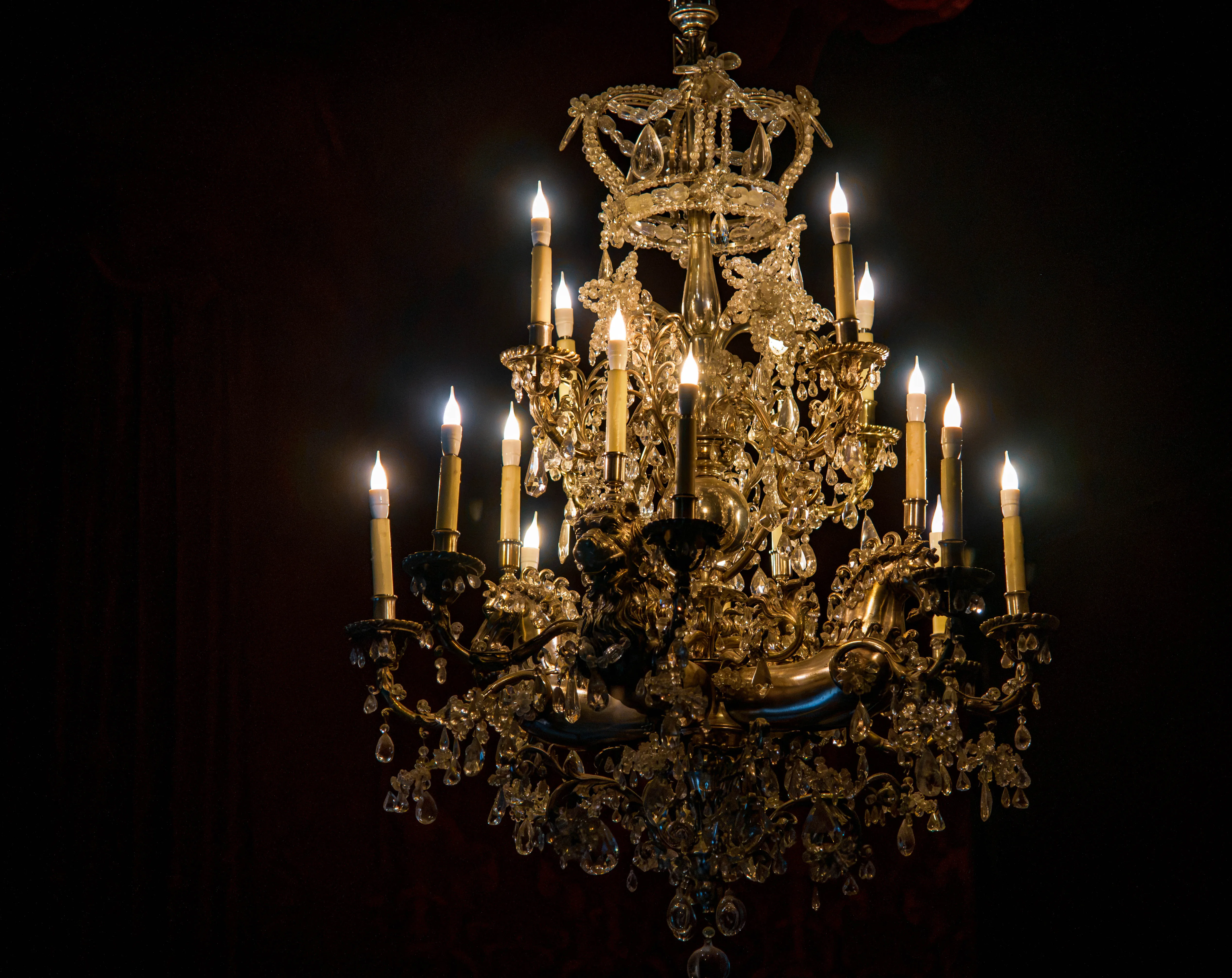 Ornate chandelier with lit candles, intricate metalwork and crystal decorations glowing against a dark background.