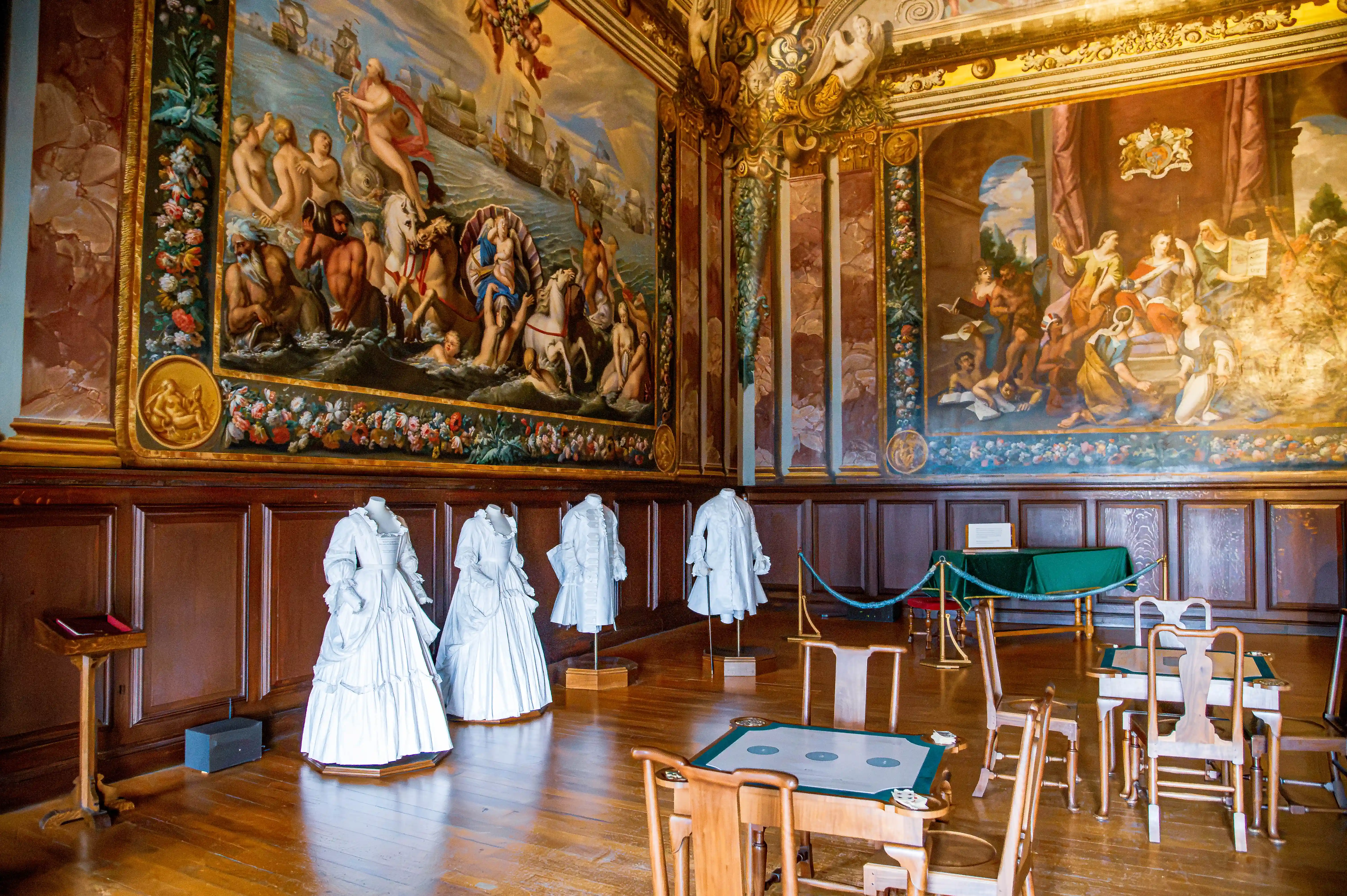 Historic room with large classical murals, white period dresses on mannequins, timber panelling and heritage furniture.