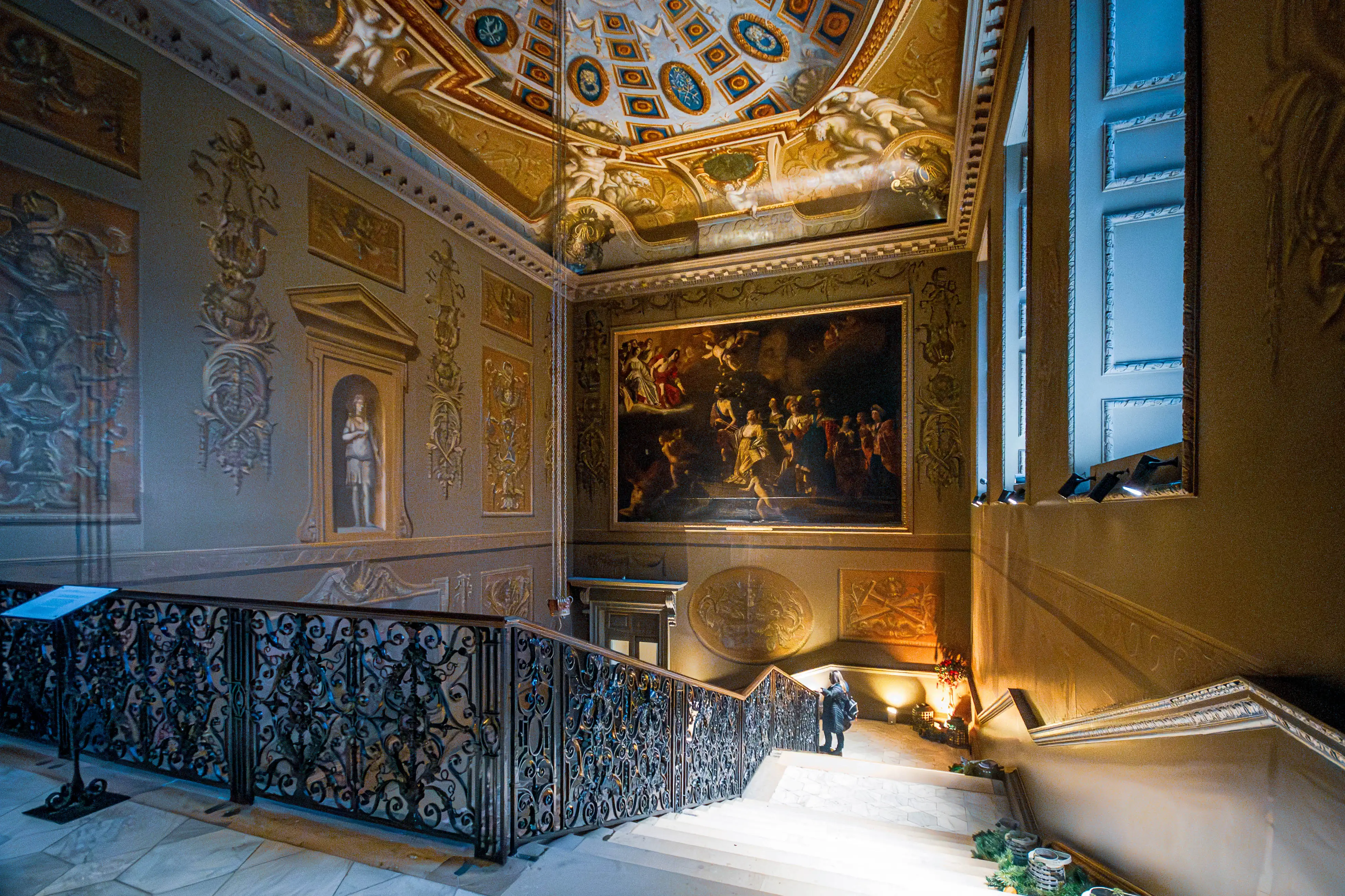 Grand staircase with marble steps, ornate iron railings, sculpted reliefs, framed artwork and Baroque ceiling fresco.