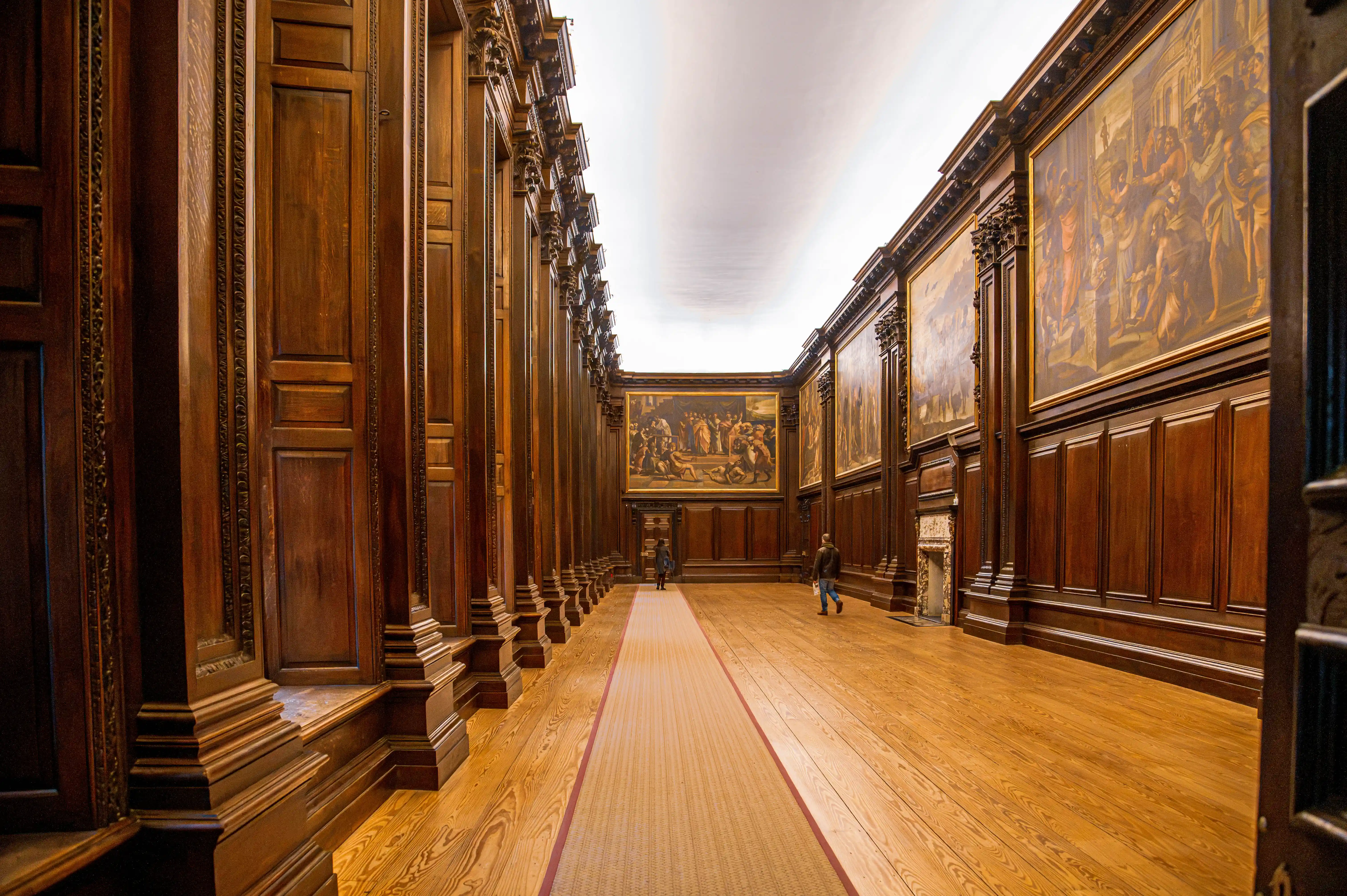 Long historic hallway with timber panelling, large framed murals and polished wooden floor with carpet runner.