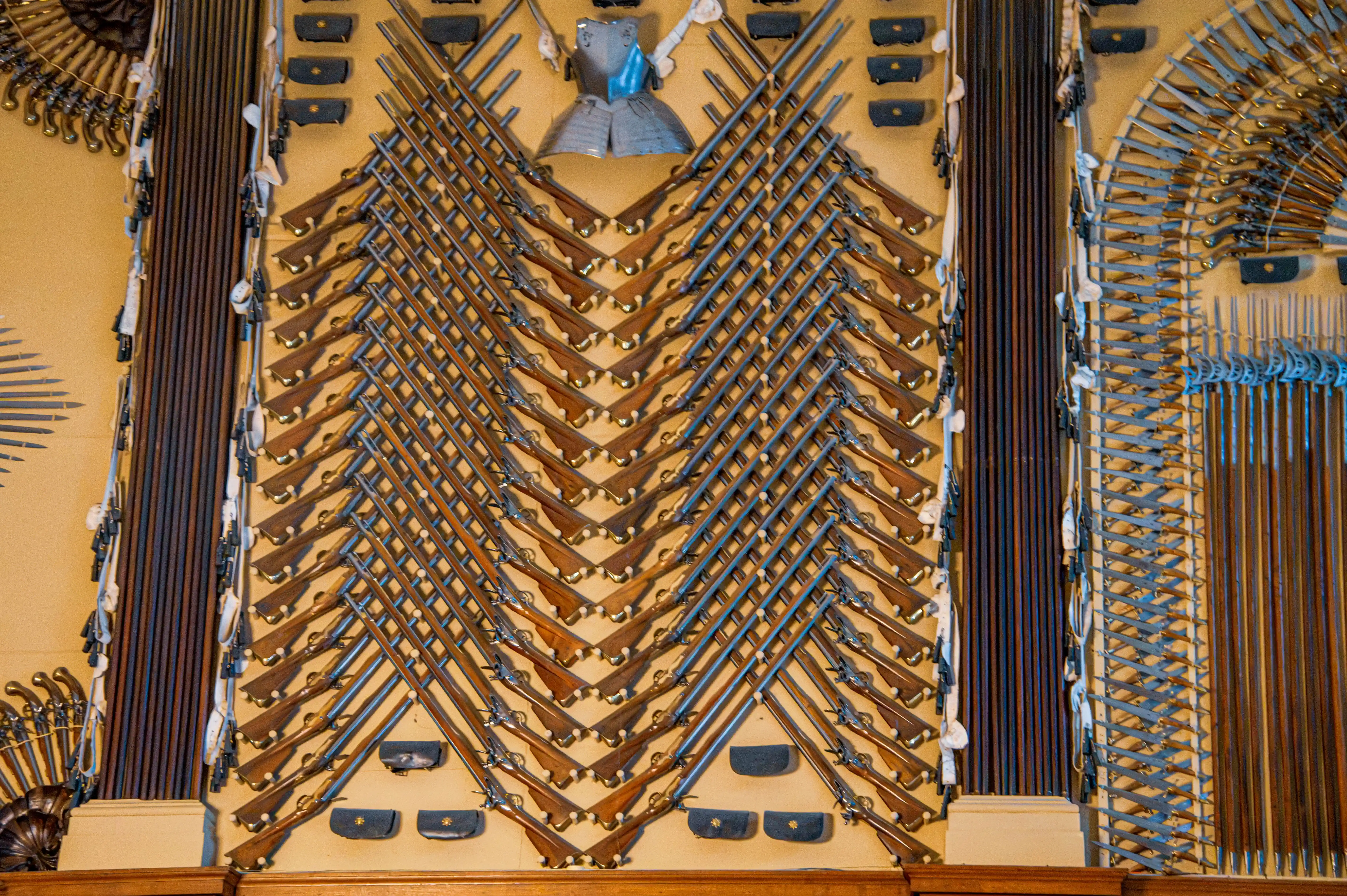 Symmetrical wall display of muskets, swords and mounted armour arranged in geometric patterns.