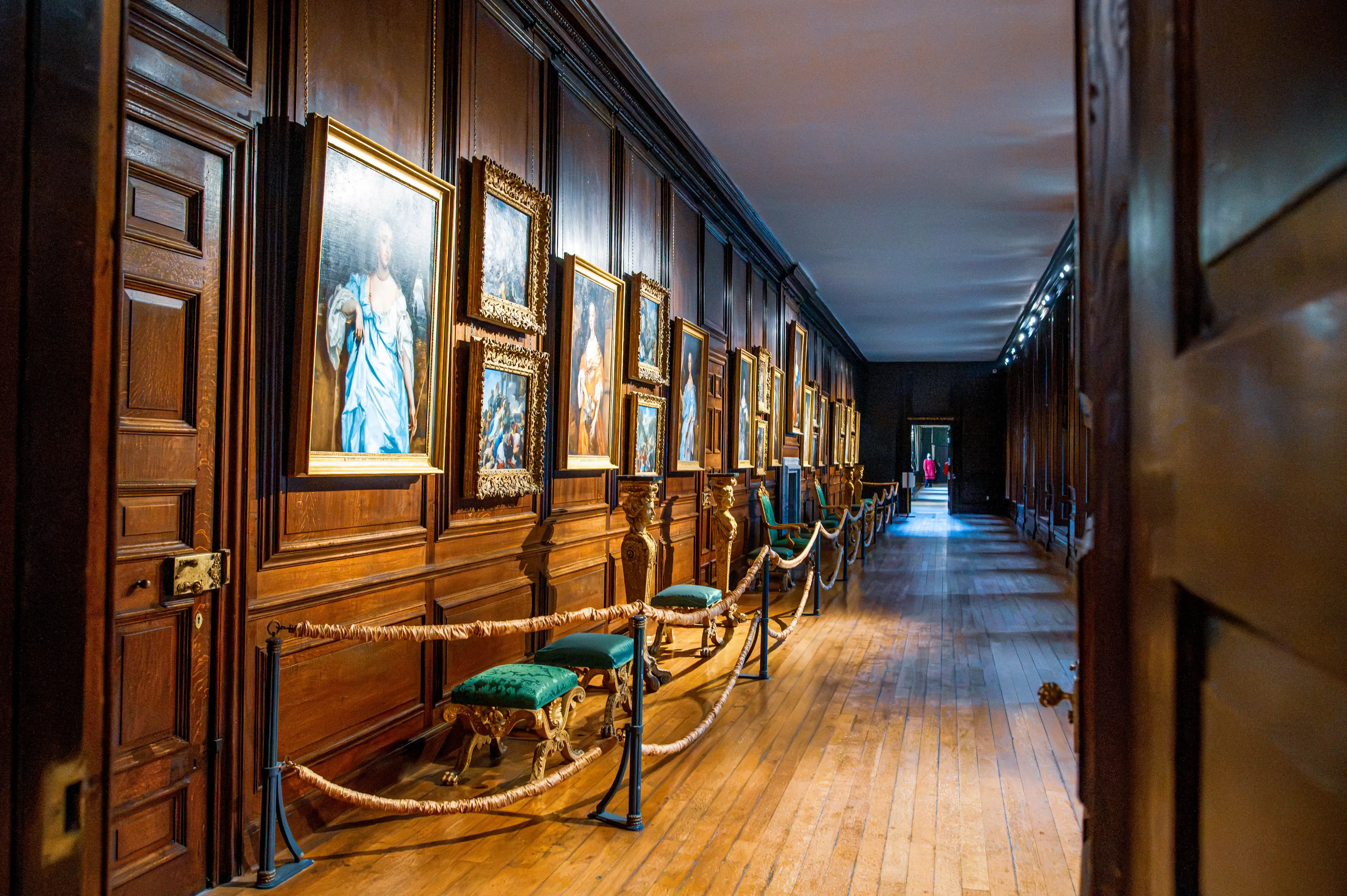 Long historic corridor with timber panelling, framed portraits and ornate chairs behind rope barriers.