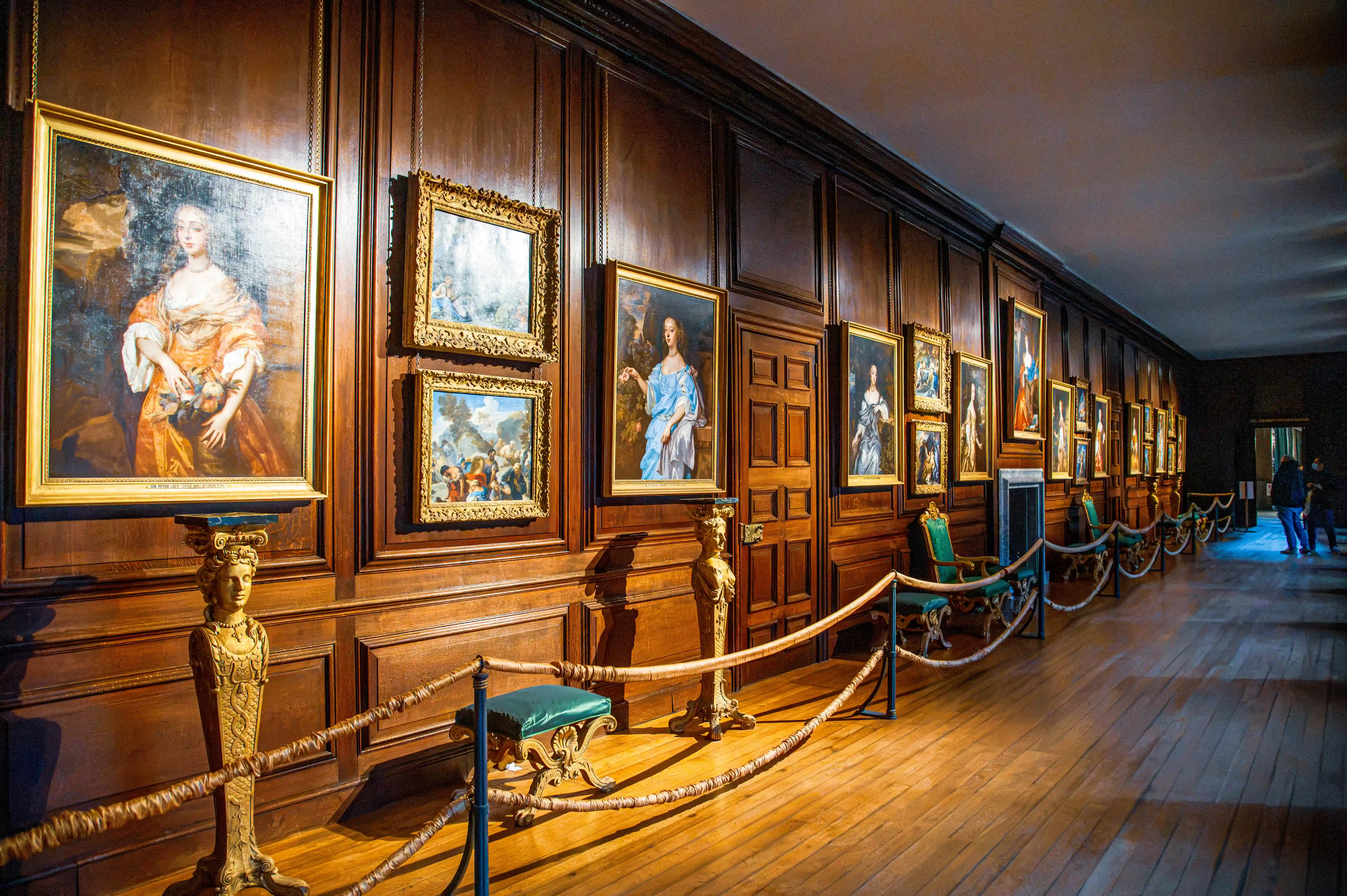 Long portrait gallery with timber panelling, gold-framed paintings, rope barriers and polished wooden floor.