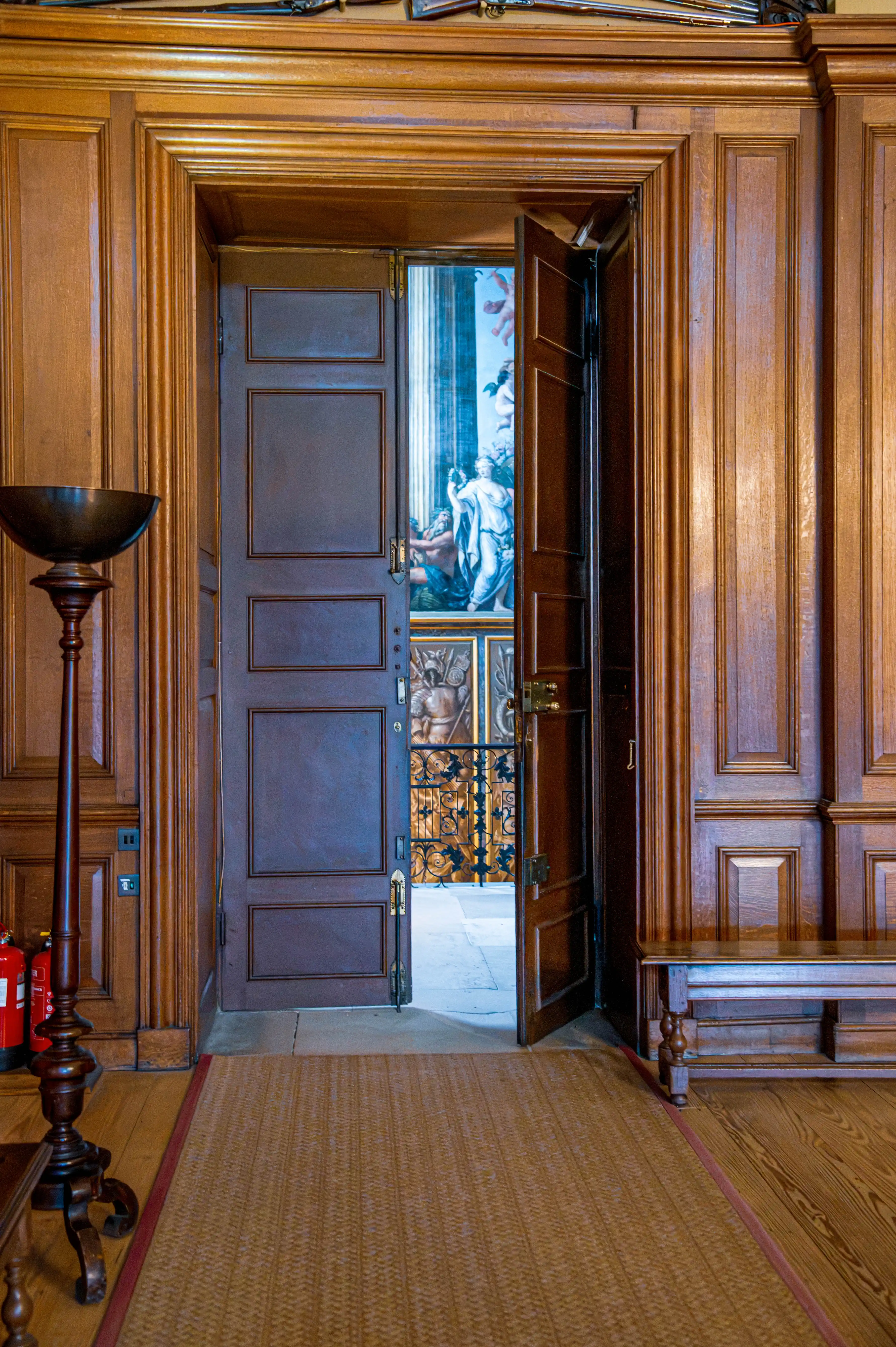 Timber-panelled room with carved woodwork, woven rug and doorway revealing a classical mural in the next chamber.