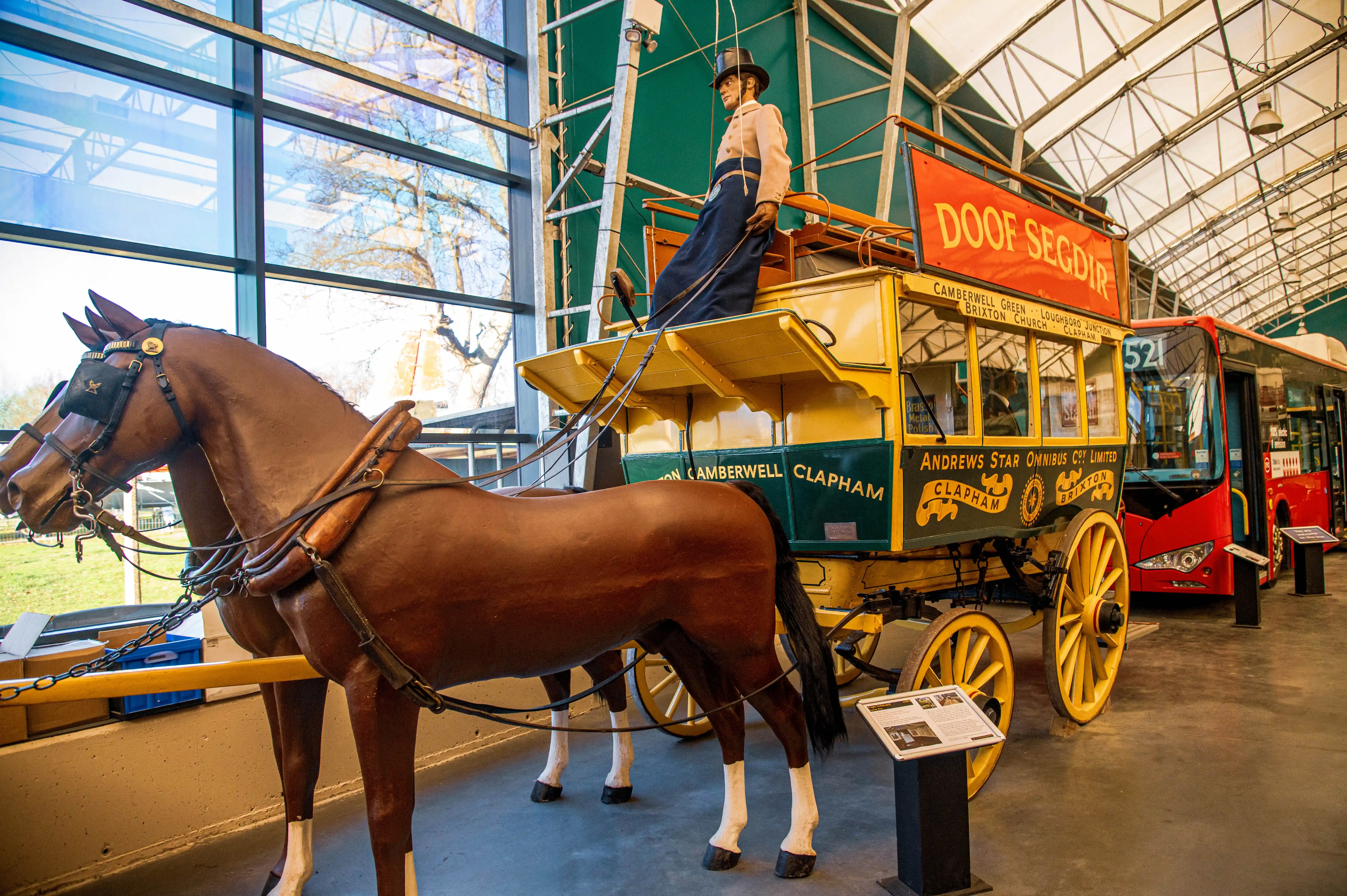 A museum exhibit showcasing a detailed model of a horse-drawn omnibus with two brown horses, standing next to a modern red bus. The omnibus has a driver mannequin on top and lettering that includes Andrews Star Omnibus Co. Ltd. and destinations like Camberwell Green and Clapham.
