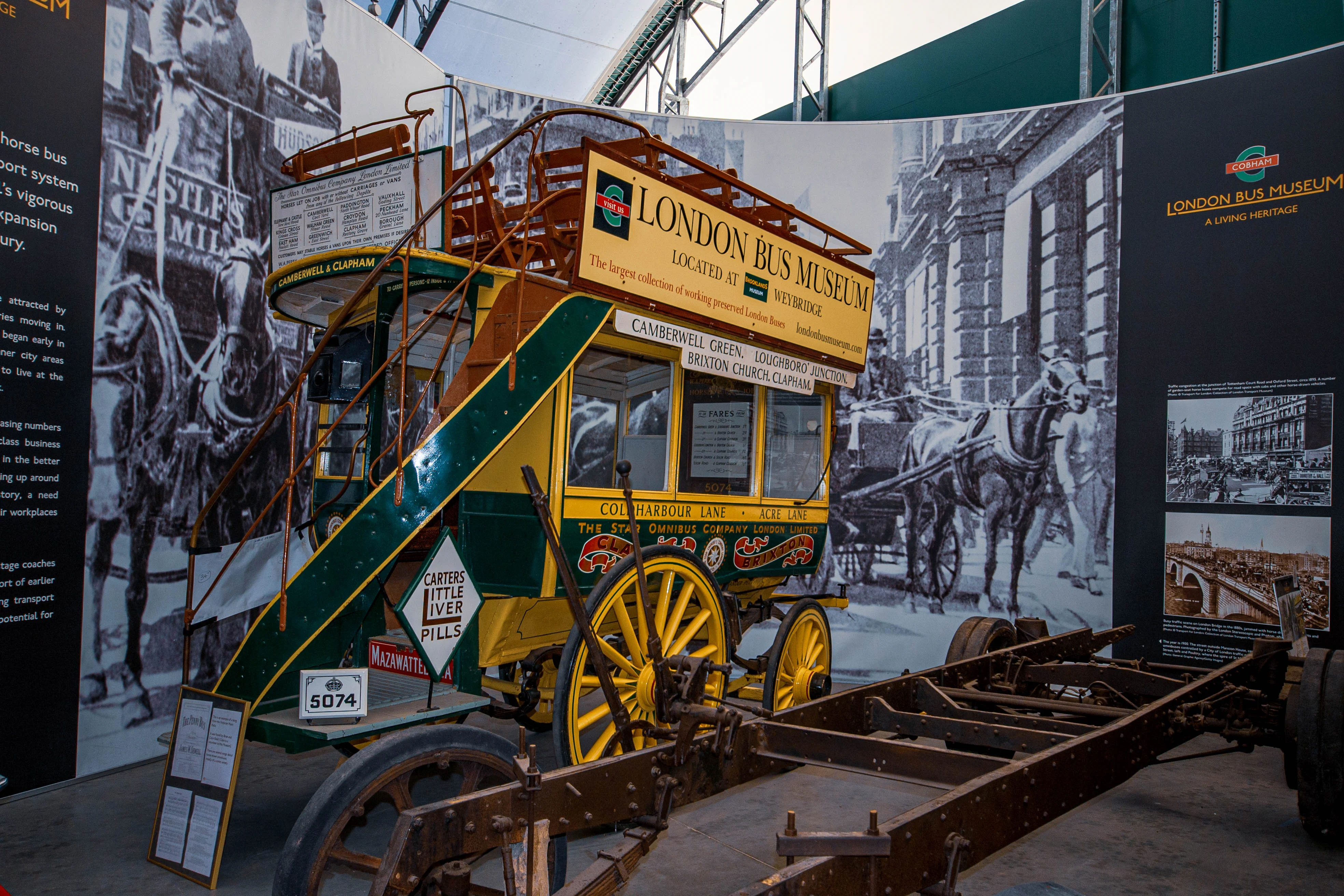 A vintage yellow and green horse-drawn double-decker bus, with an open-top upper deck and ornate stairs, stands on display in a museum. Historical murals of London street scenes with horse-drawn vehicles are visible in the background, alongside the London Bus Museum logo. The chassis of a vehicle is in the foreground.