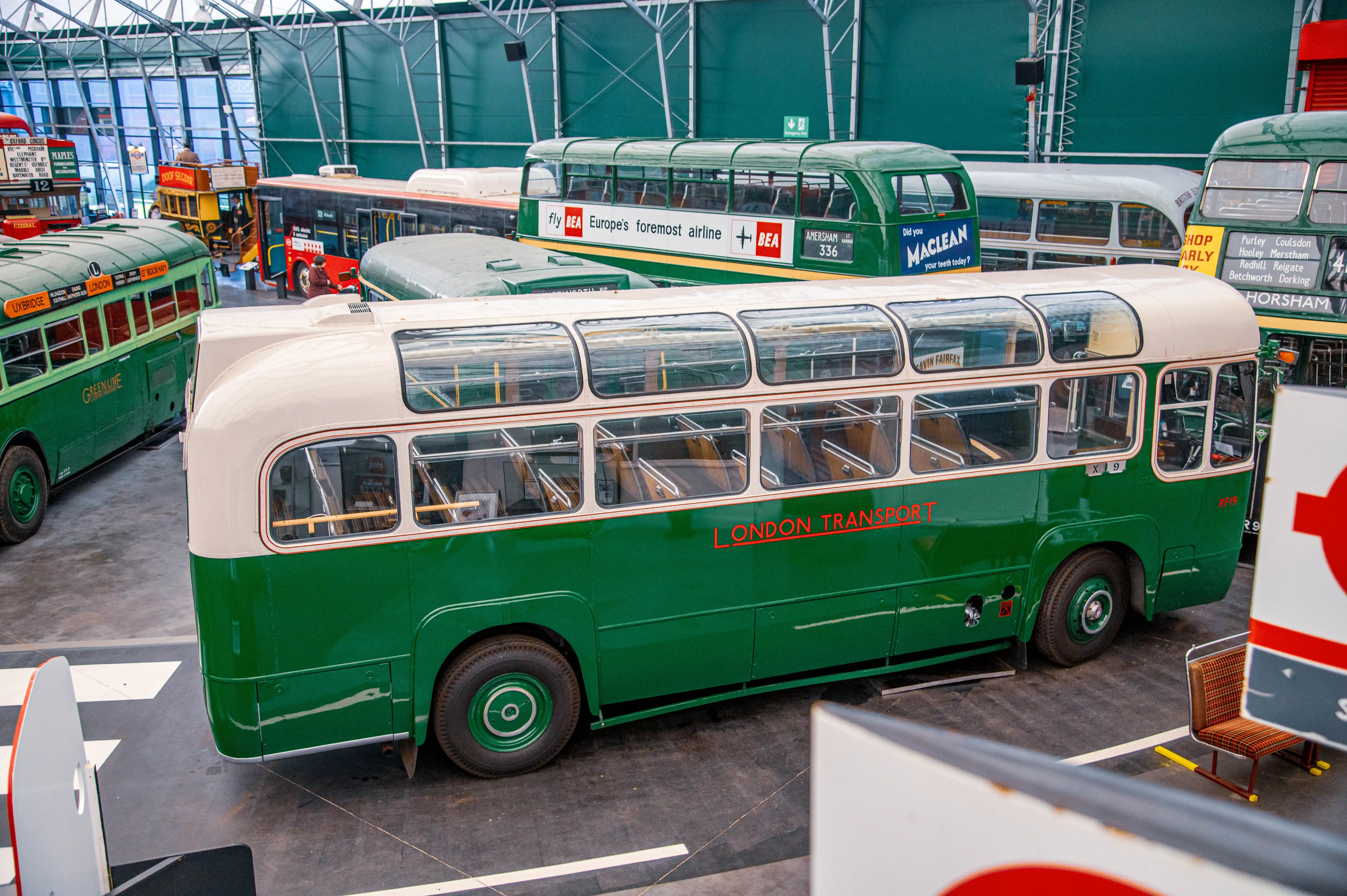 A close-up, high-angle view of a vintage green and cream London Transport bus parked indoors. The bus has large windows, chrome detailing, and the distinctive London Transport branding in red. Other vintage buses and vehicles are visible in the background.