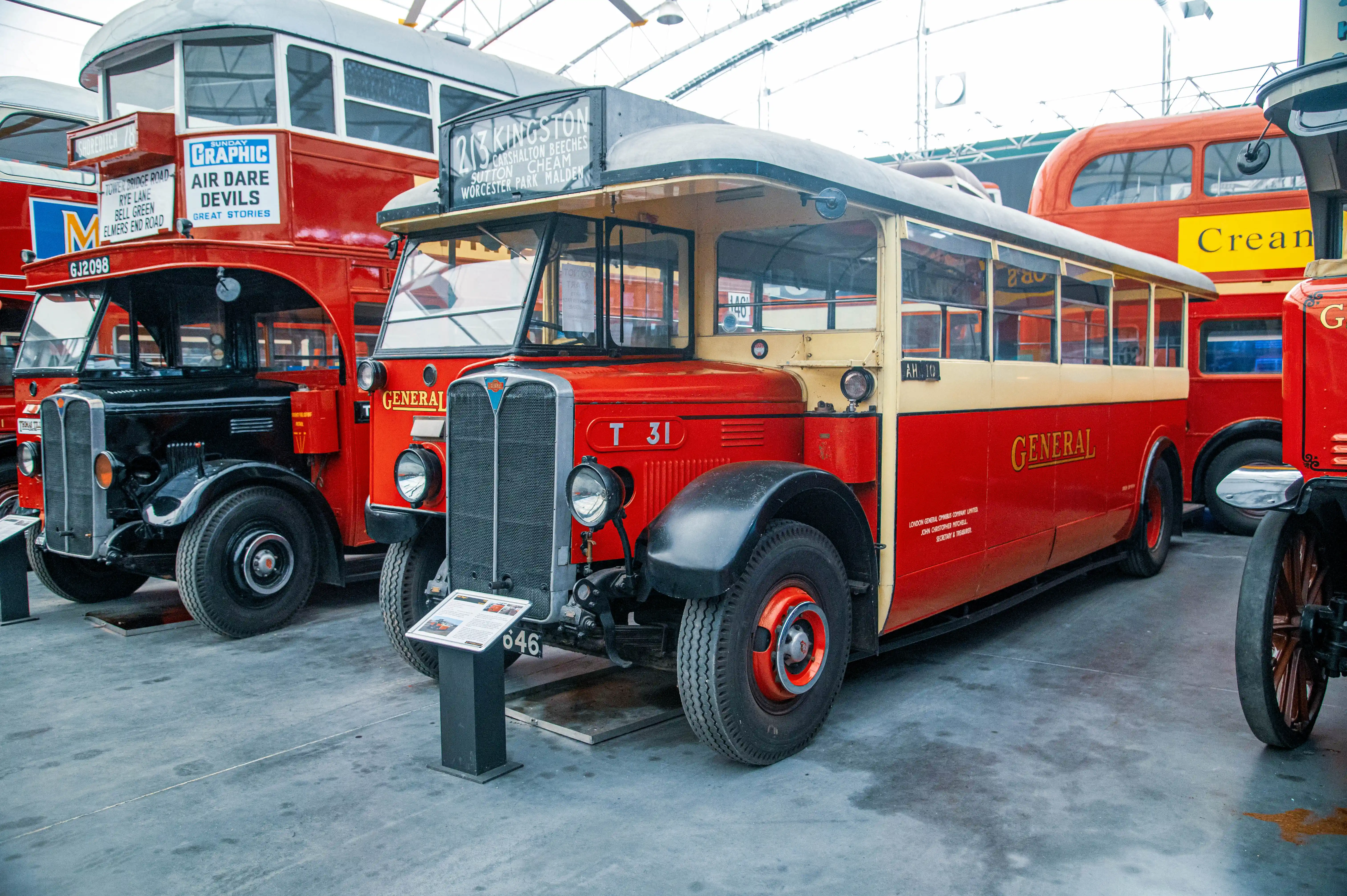 Three vintage buses, two red and one with a black front and red body, are parked side-by-side in an exhibition hall. The bus in the centre is predominantly red with cream accents and the word GENERAL in gold lettering. A route sign above its windscreen reads 213 KINGSTON.